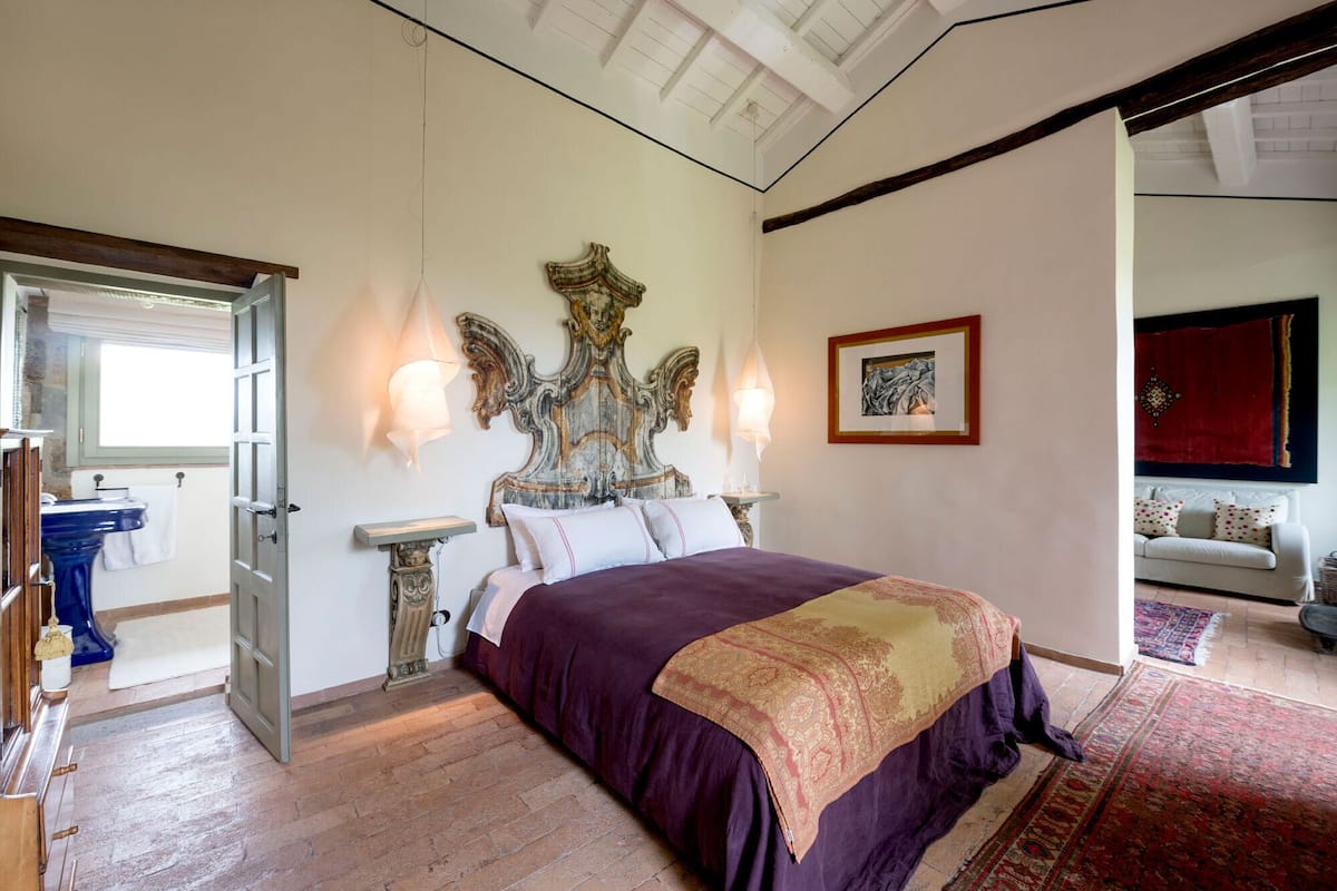 A bedroom with a large bed, a decorative headboard, two wall-mounted lamps, a framed artwork, and a view of a living area with a sofa and a red curtain.