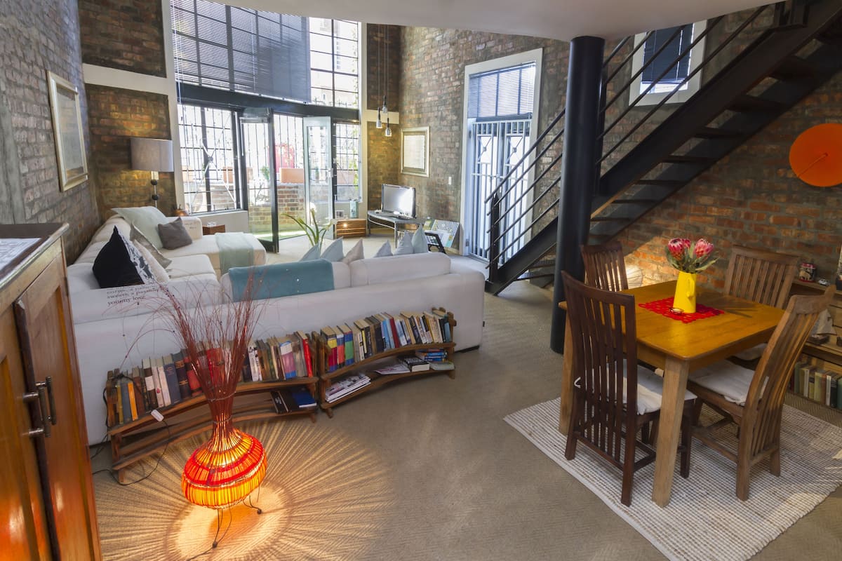 A spacious living room with a white sofa, a wooden coffee table, and a bookshelf filled with books. A staircase is visible in the background.