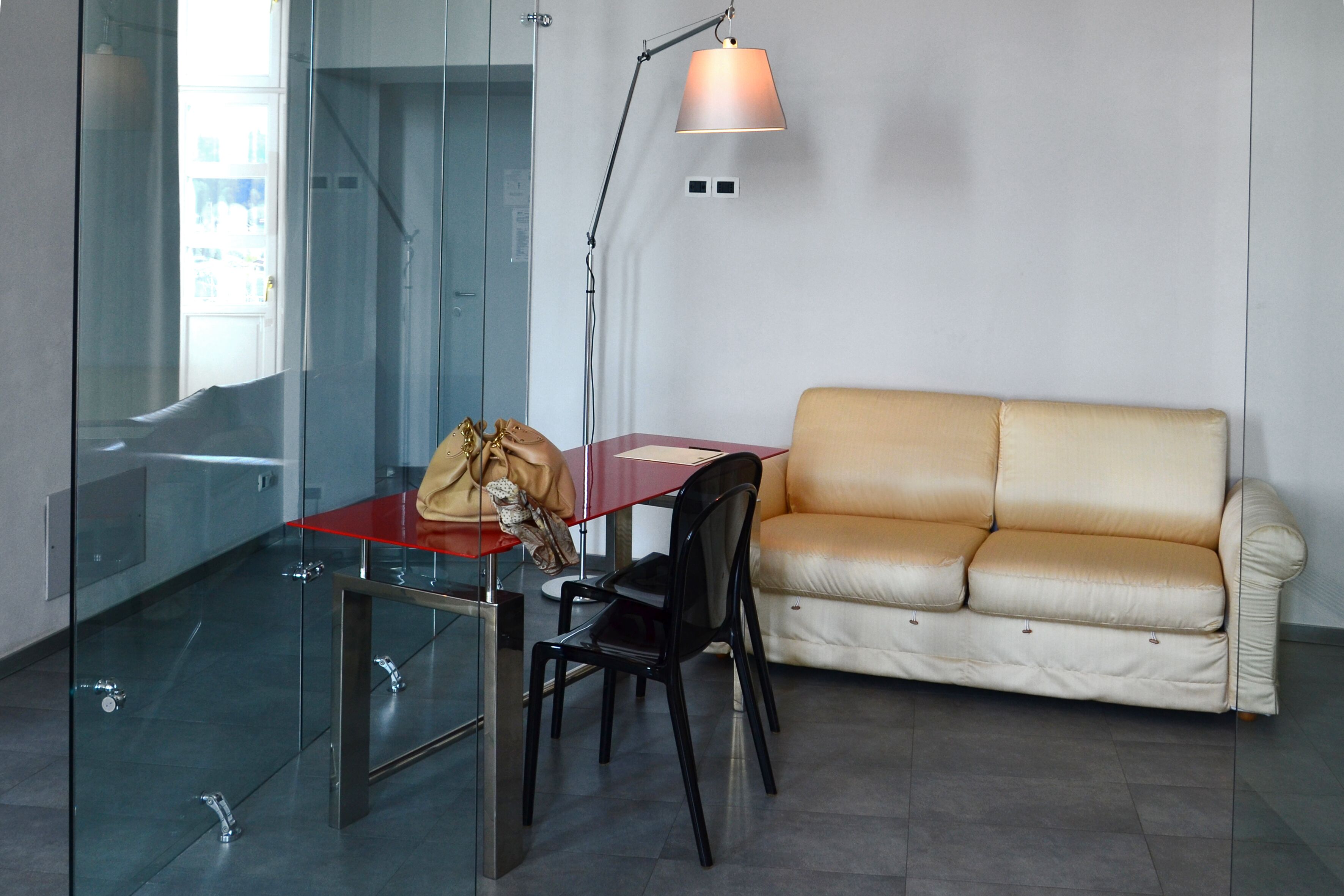 A modern room with a glass partition, a red table, a black chair, a beige sofa, and a floor lamp.