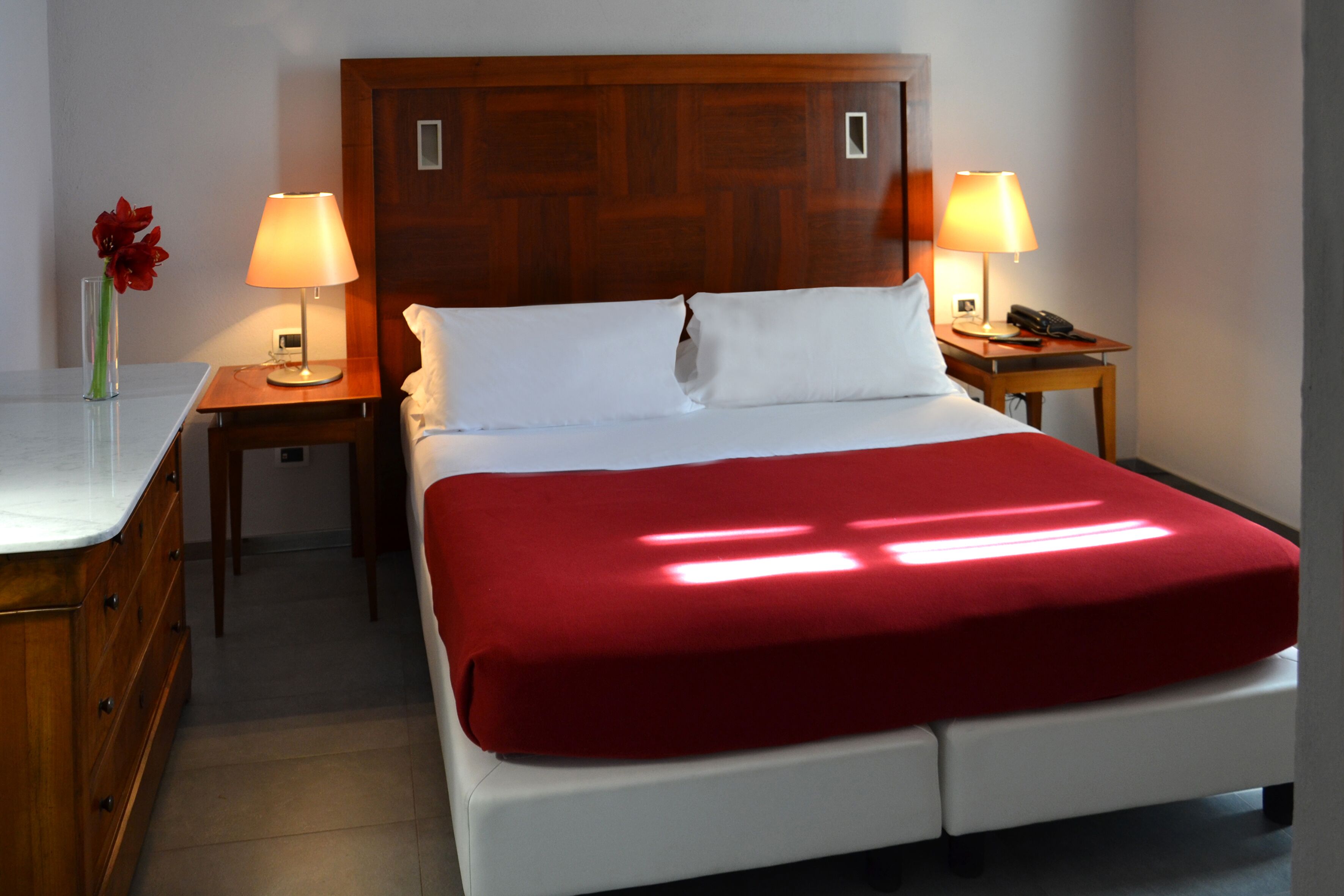 A hotel room with a wooden headboard, two bedside tables with lamps, a red bedspread, and a vase with flowers on the nightstand.