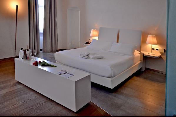 A modern hotel room with a large bed, bedside lamps, a white bedside table with a bottle and glass, and a view of the outdoors through the window. - Castello di Montaldo Torino (Montaldo Torinese)
