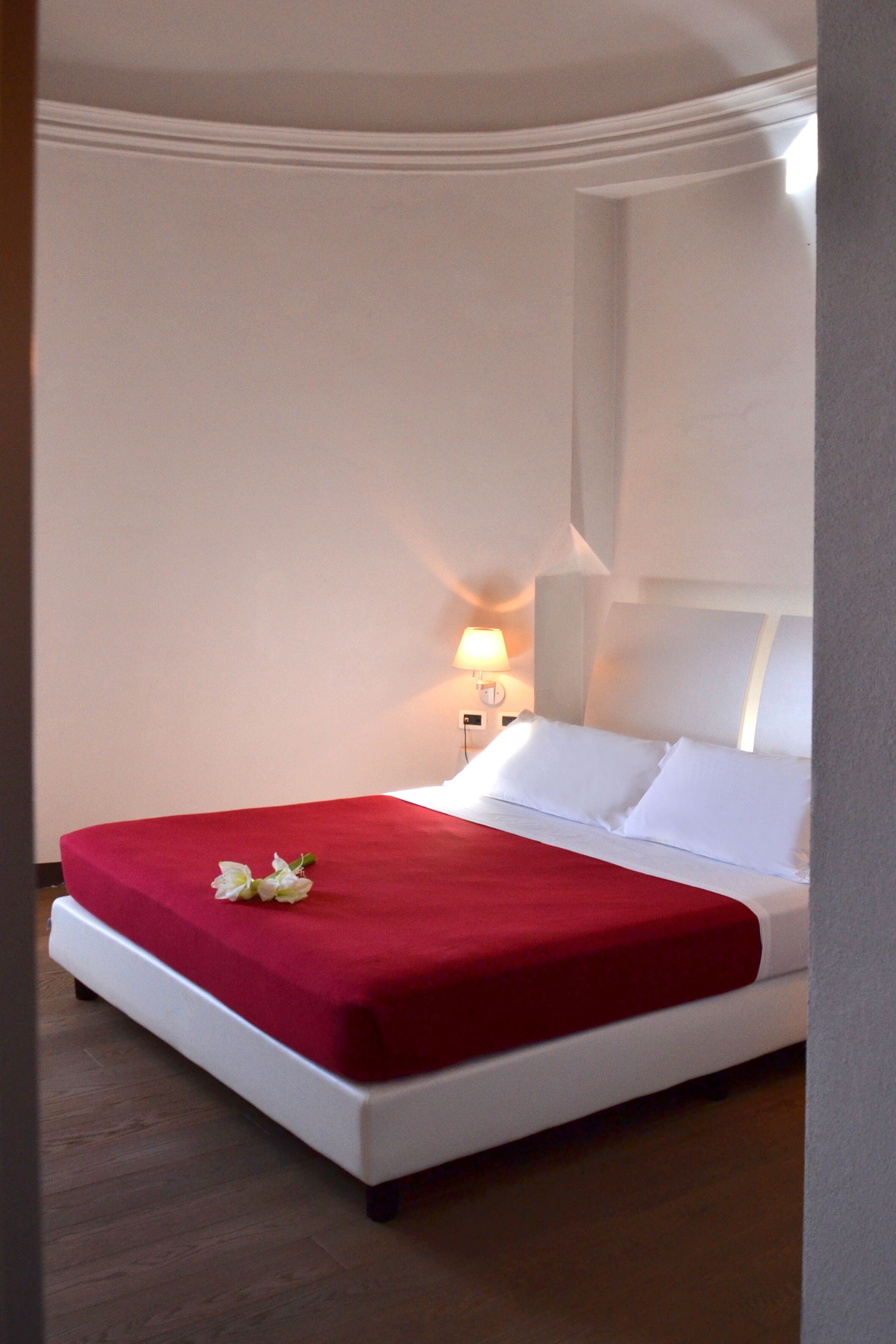 Un letto rifatto con una coperta rossa e cuscini bianchi, una lampada da comodino e un portalampada a parete.