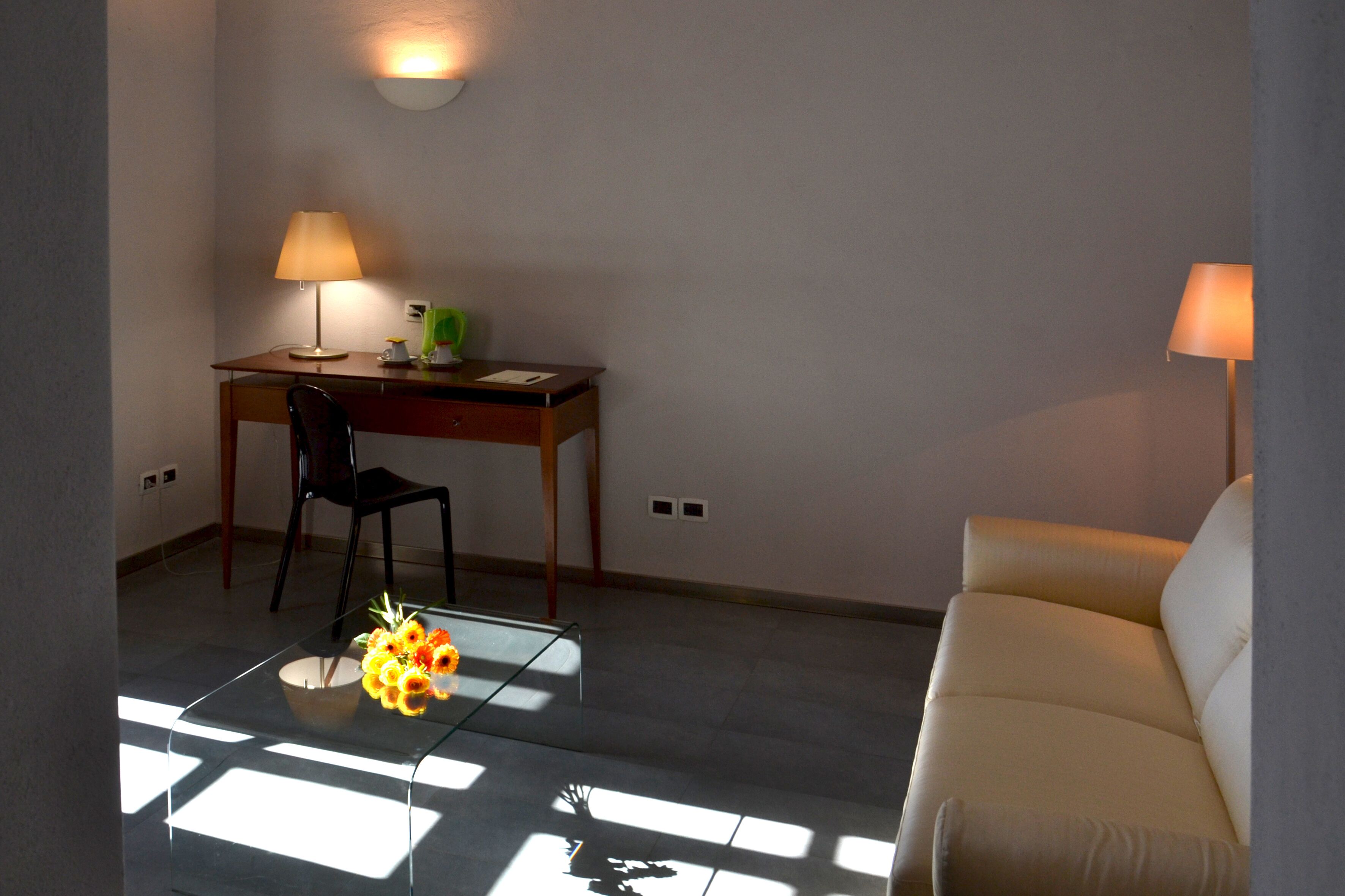 A room with a glass coffee table, a desk with a chair, a sofa, and two wall-mounted lamps.