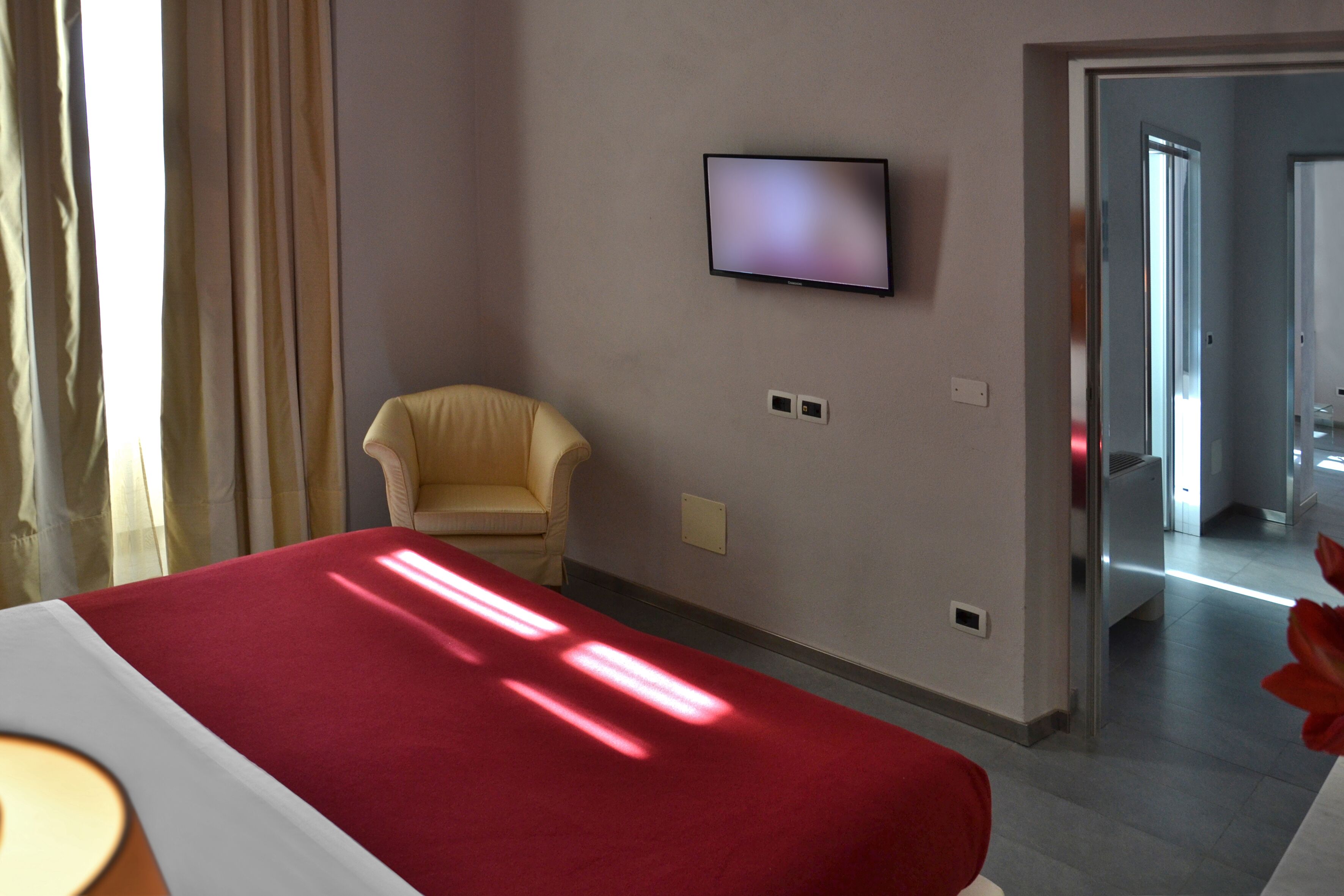 A hotel room with a bed, a chair, a TV mounted on the wall, and a door leading to another room.