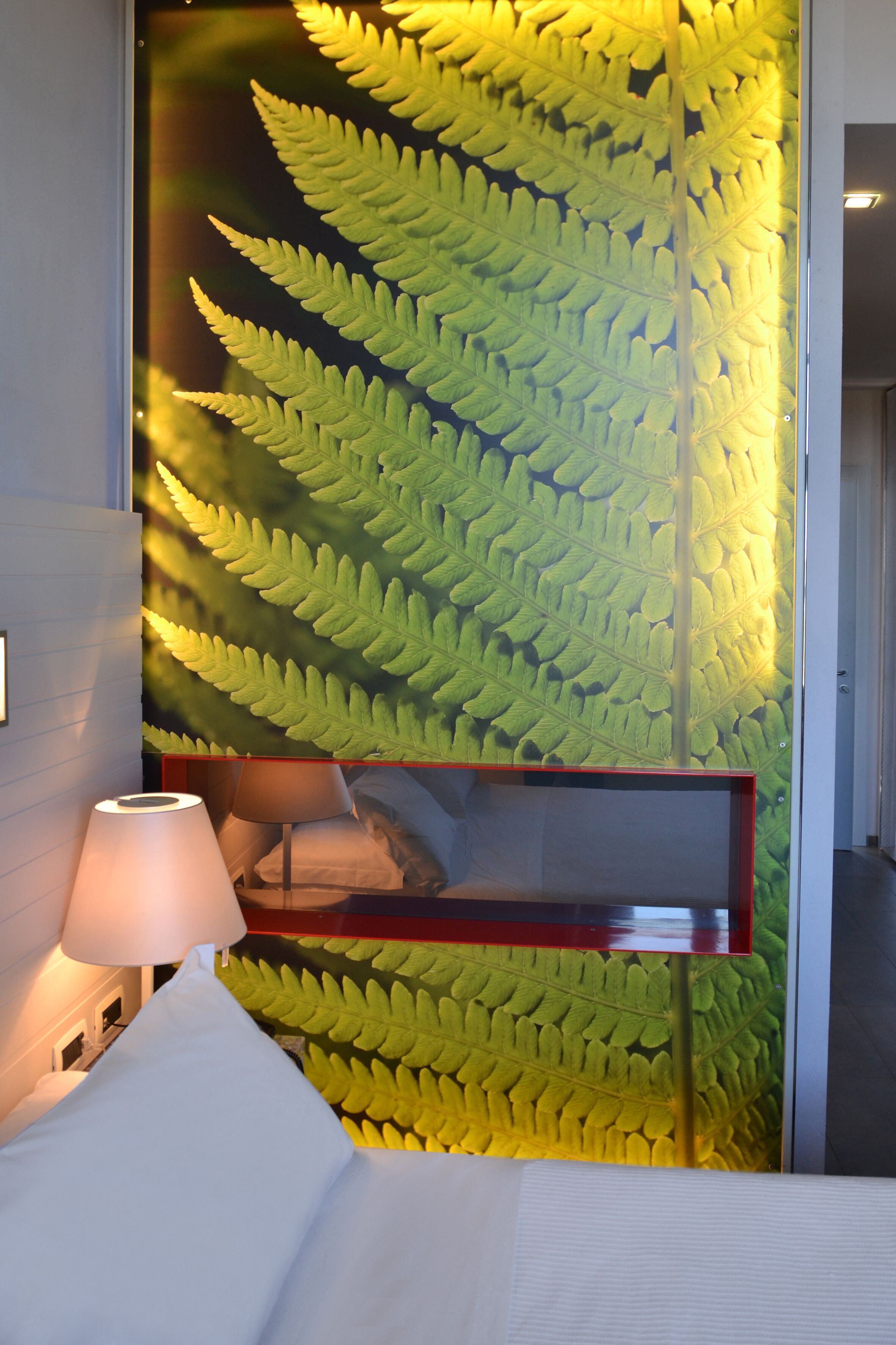 A room with a large fern leaf wall, a bed with white linens, and a bedside lamp.