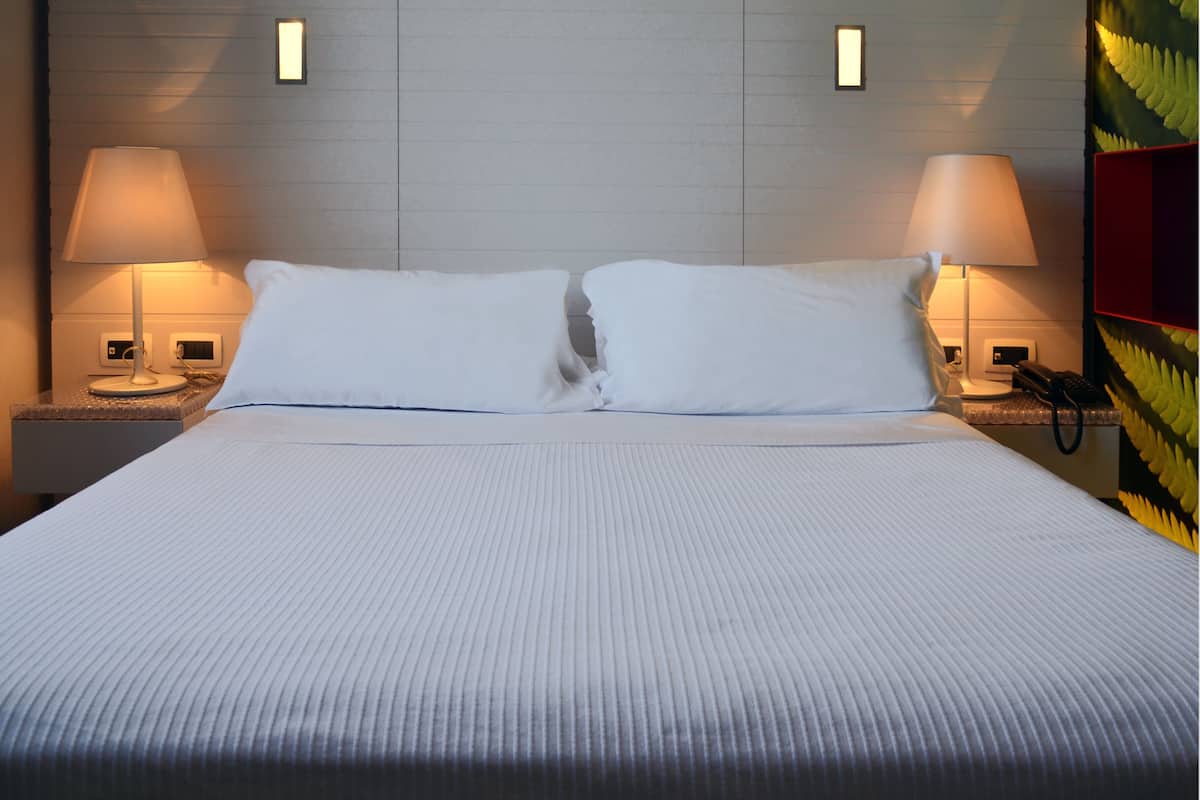 Een net opgemaakt bed met witte lakens, twee bedlampen en een nachtkastje met een telefoon.