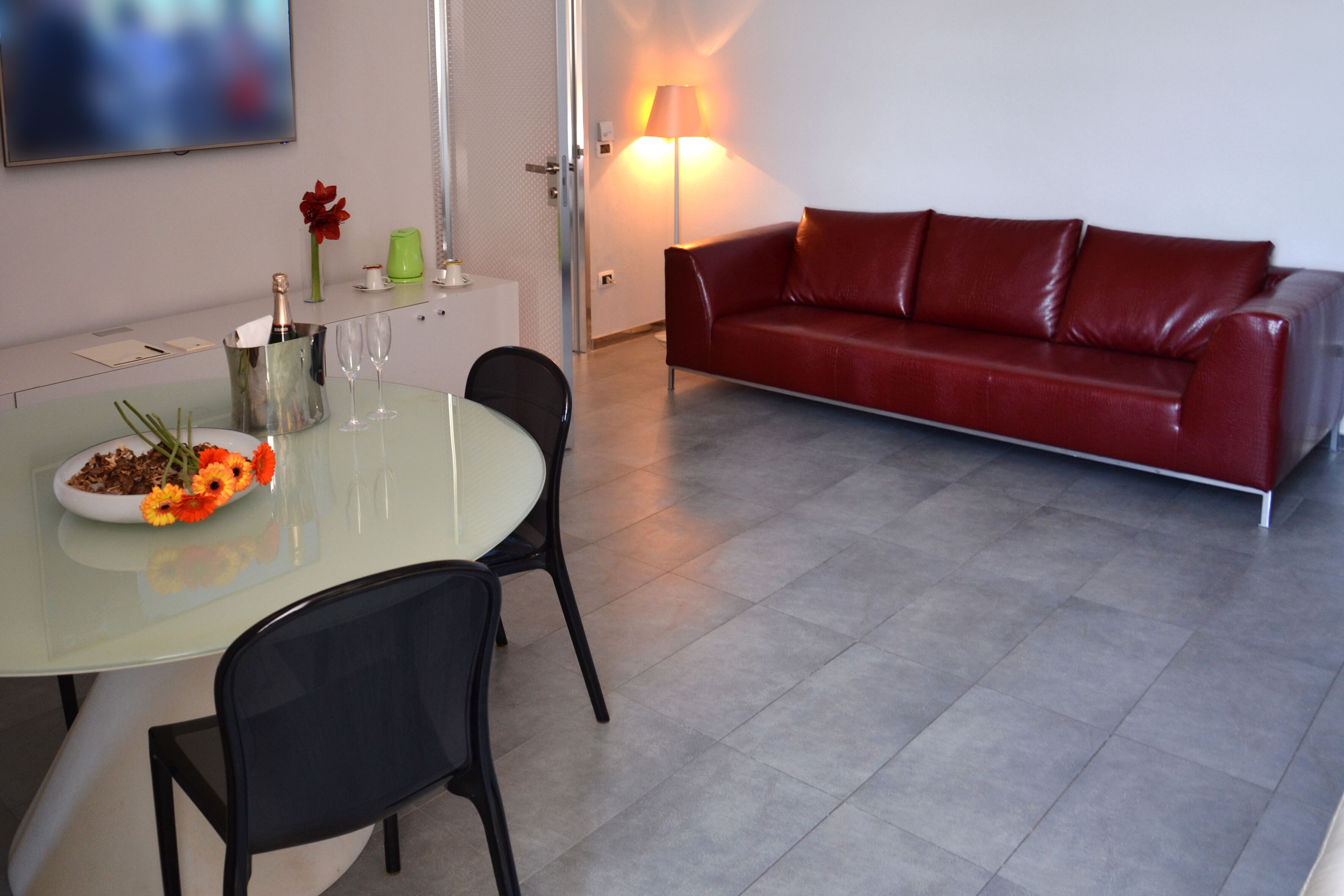 A modern living room with a glass dining table, two black chairs, a red leather sofa, a flat-screen TV mounted on the wall, and a floor lamp.