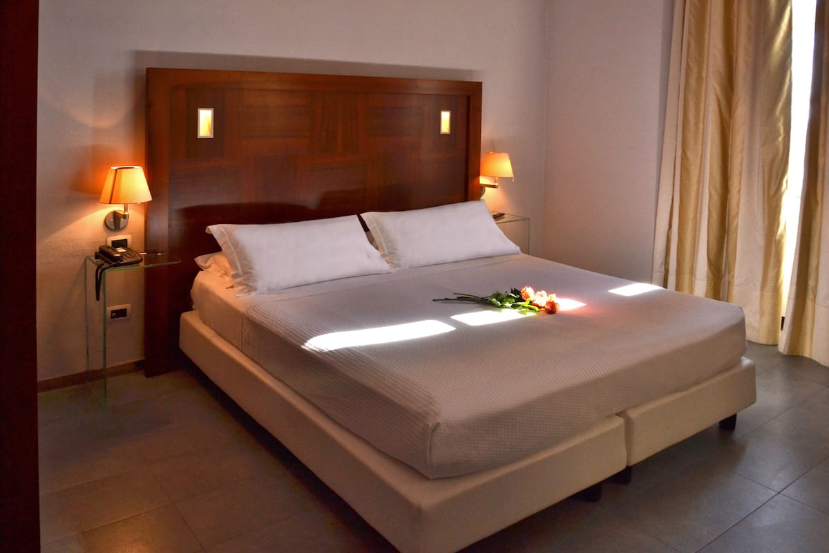Een net opgemaakt bed met een houten hoofdbord, twee bedlampjes en een bloemstuk op het bed.