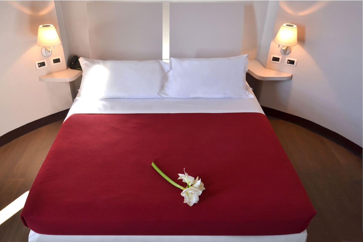 Een hotelkamer met een bed, witte kussens, een rode spreideken en twee bedlampen.