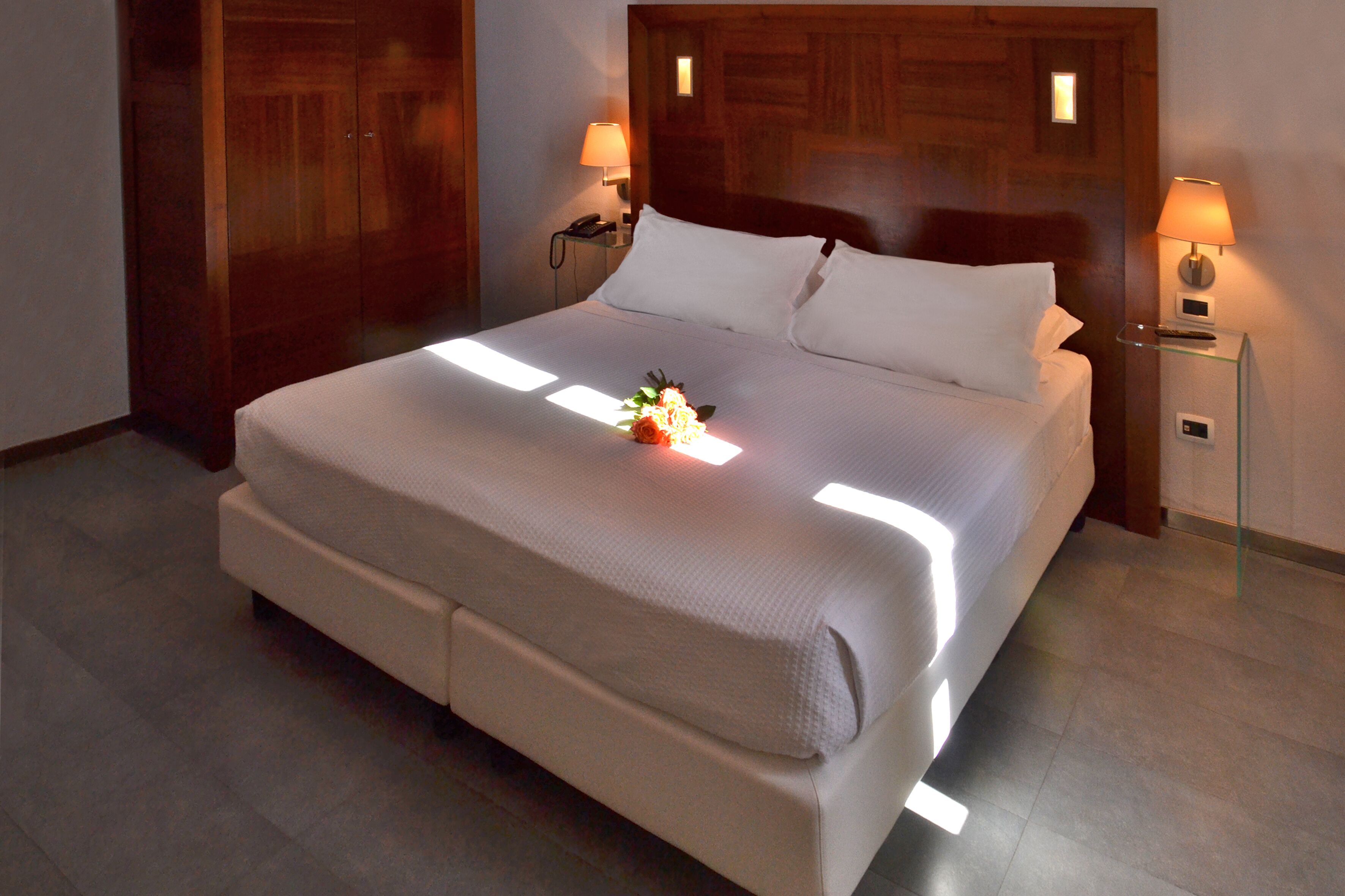 Een opgemaakt bed met een witte dekbedovertrek en kussens, twee nachtlampjes en een klein bloemenarrangement op het bed.