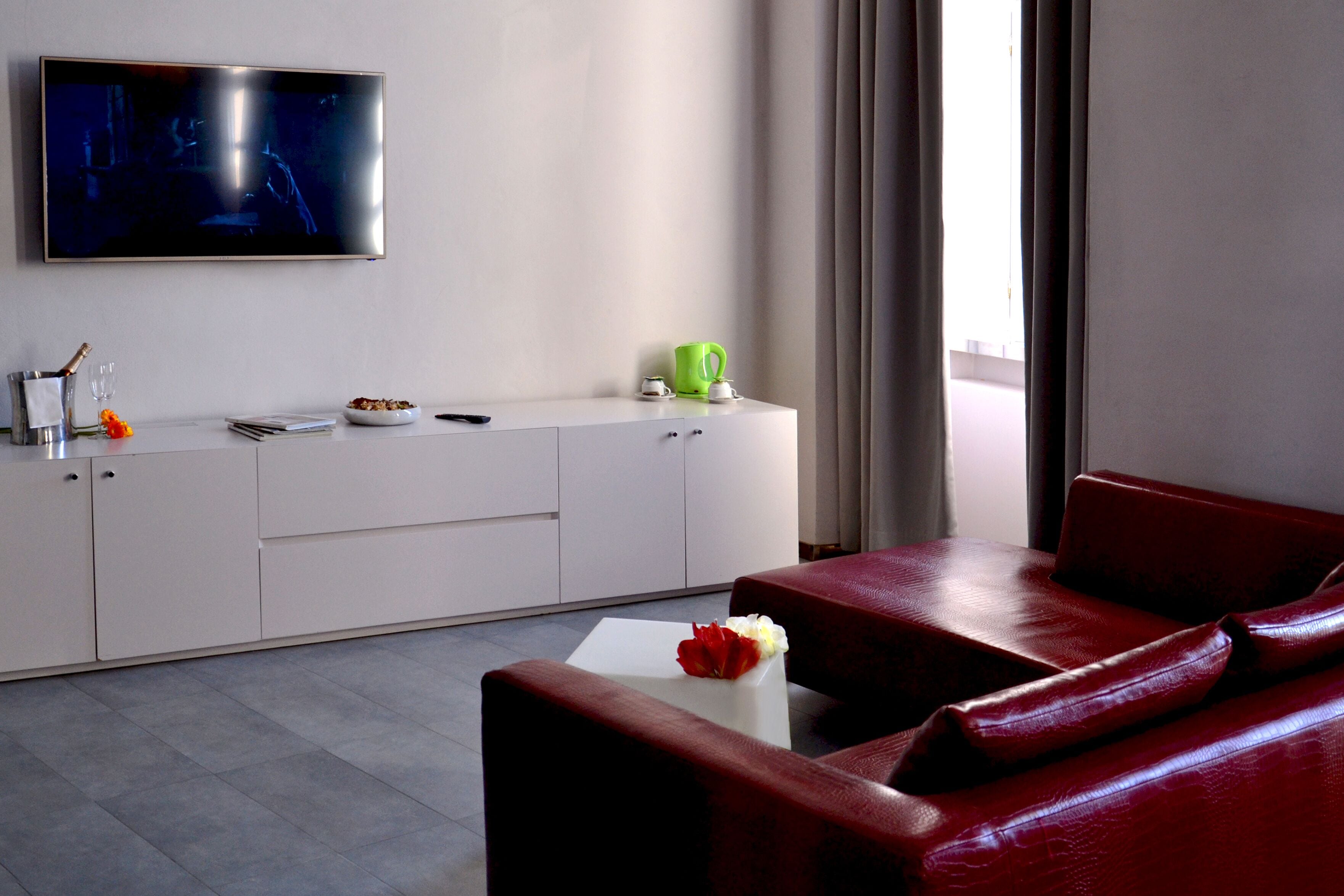 Un salon moderne avec un téléviseur à écran plat, une console blanche sur laquelle est posée une tasse verte, un canapé en cuir rouge et une petite table blanche agrémentée de fleurs.