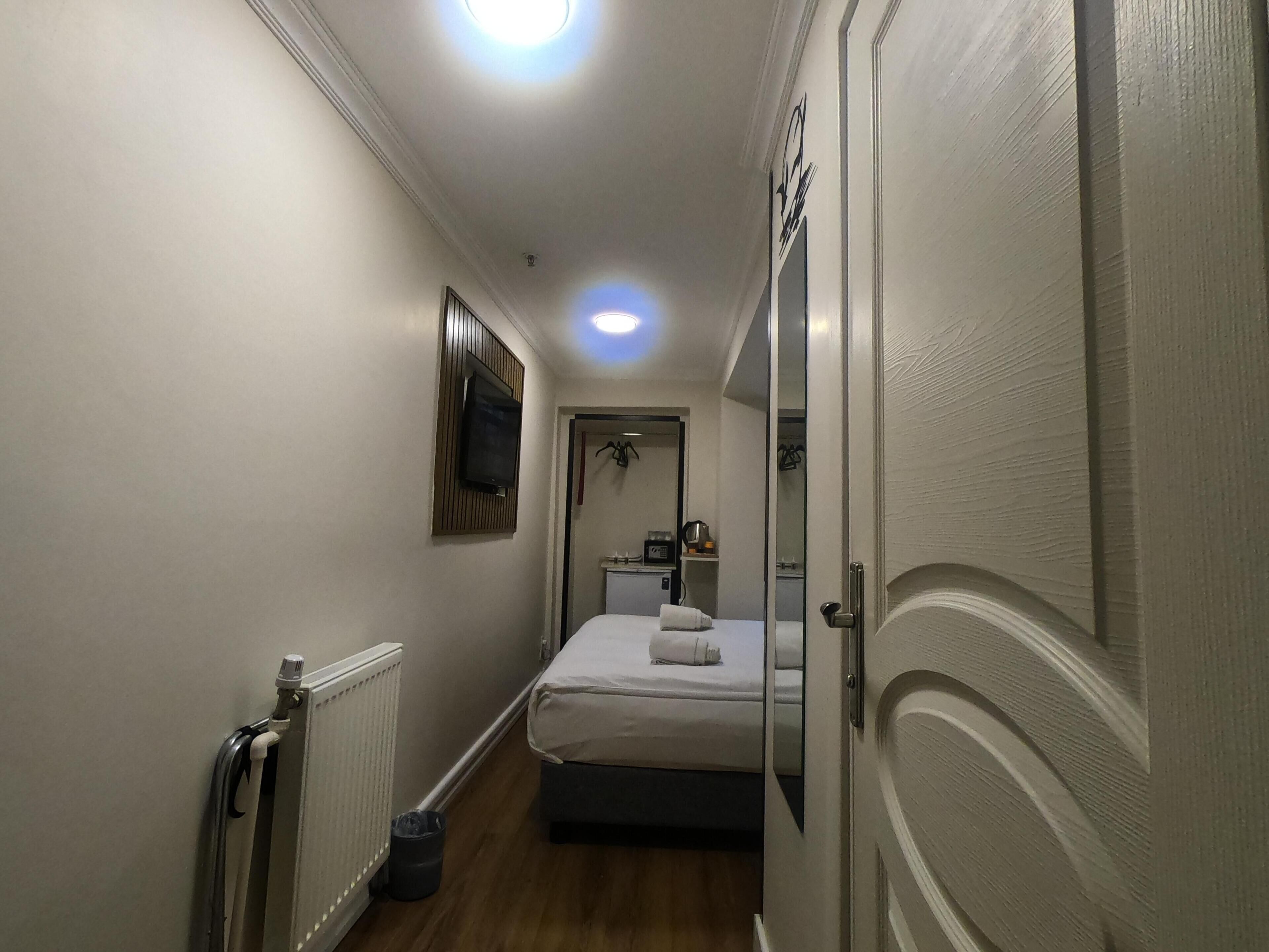 A narrow hotel room with a bed, a radiator, a small table with a kettle, and a mirror.