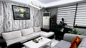 A modern living room with a grey sectional sofa, a coffee table, and a black office chair. There is a framed painting on the wall, a potted plant, and a large window with blinds. - Naila Hotel (Istanbul)