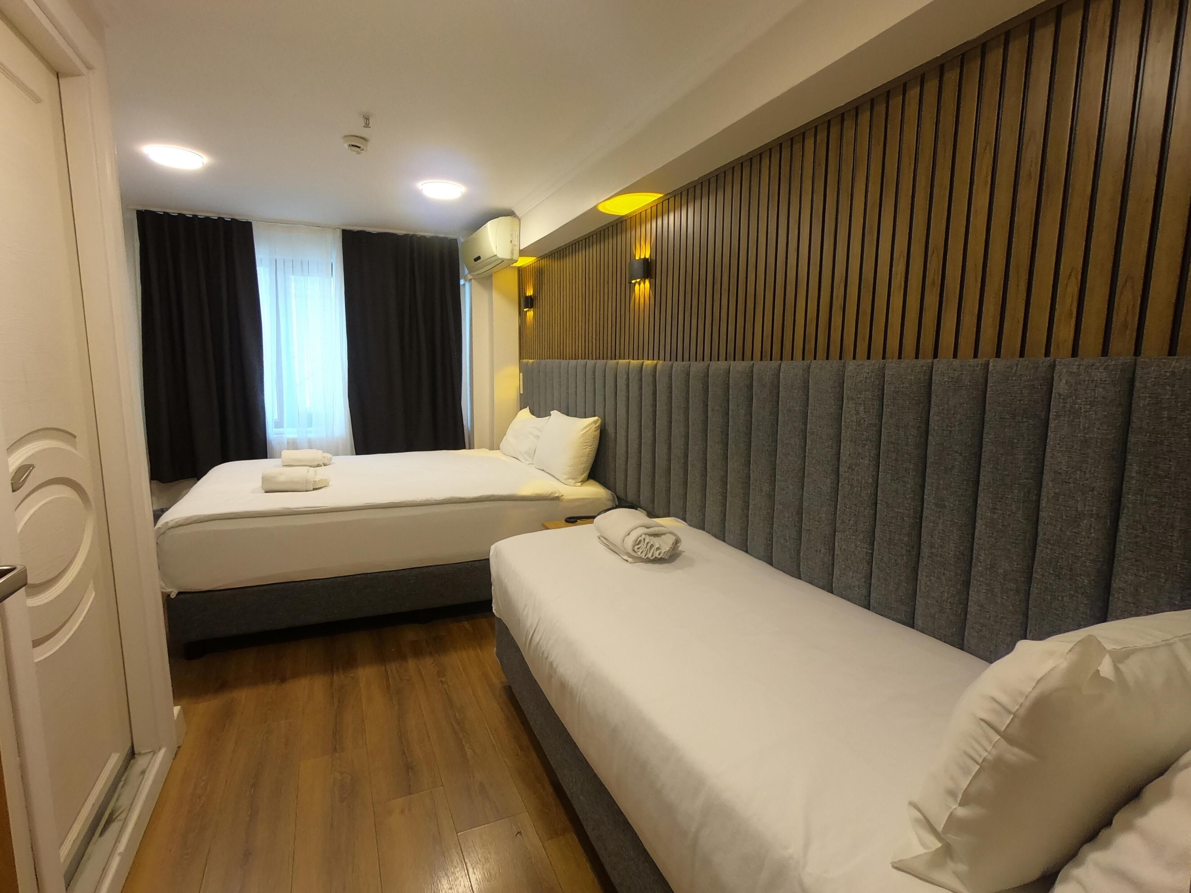 A hotel room with two beds, a wooden floor, a window with curtains, and a wall with vertical wooden slats.