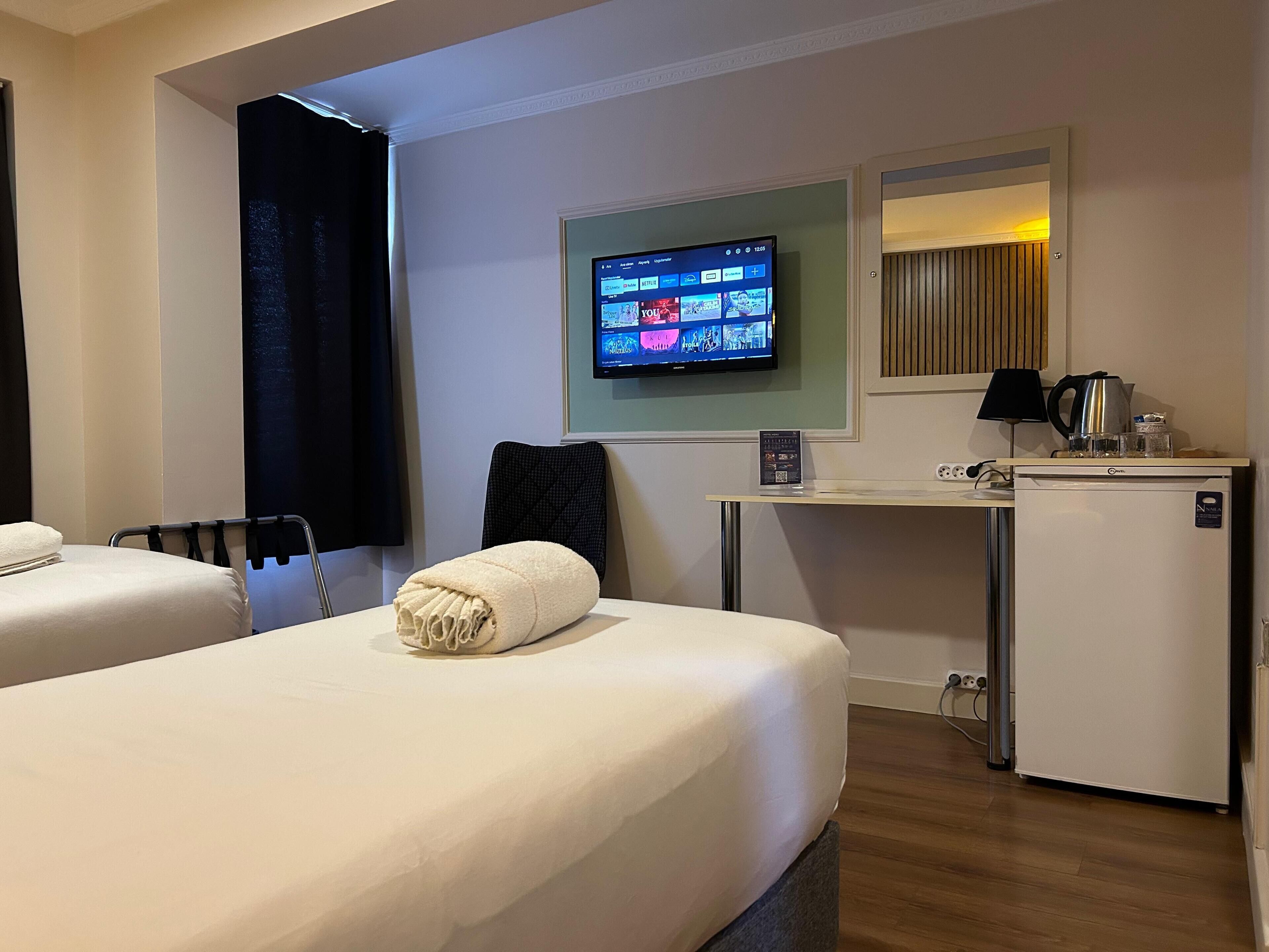 A hotel room with two beds, a TV mounted on the wall, a desk with a kettle, and a small refrigerator.