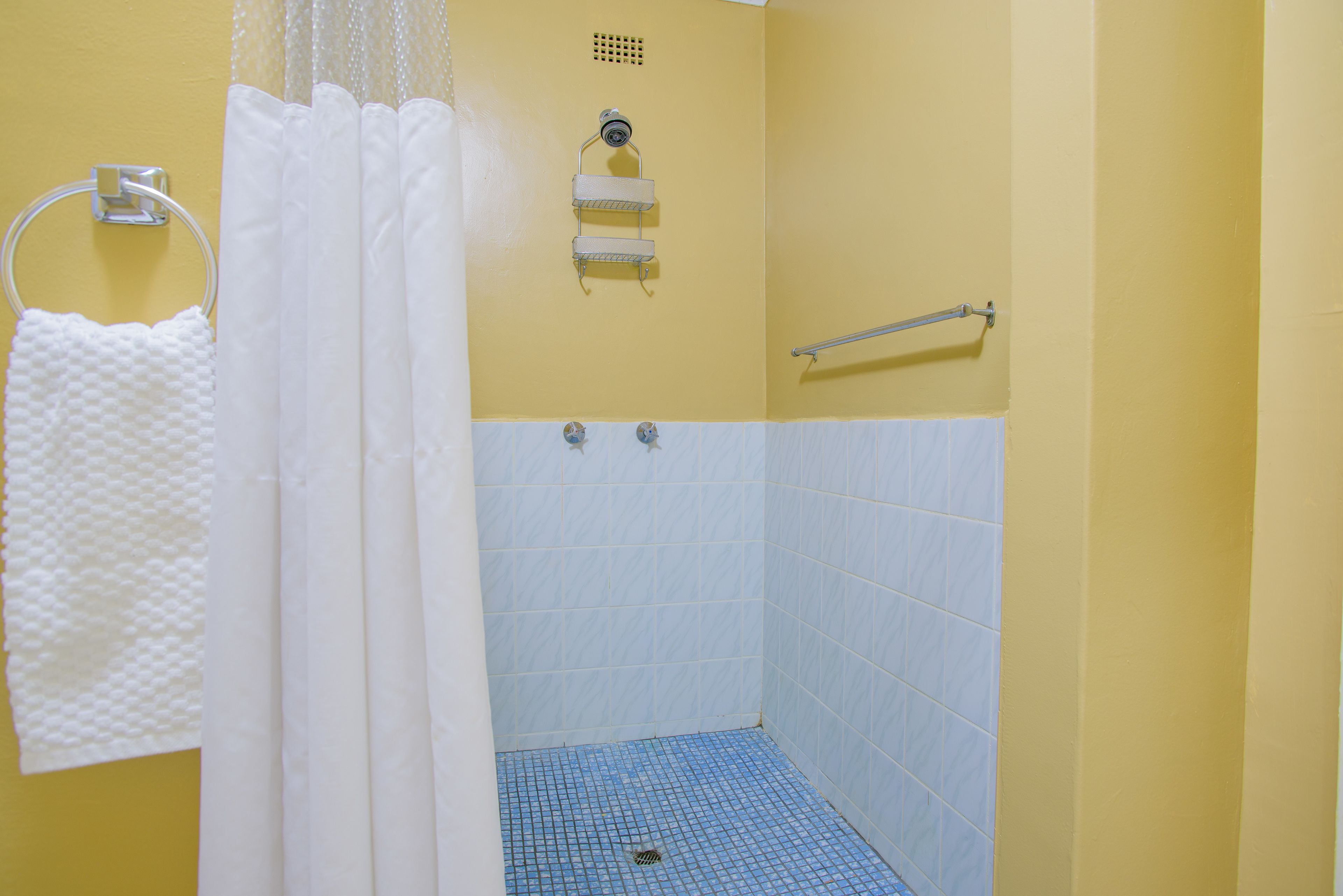 A small, yellow-walled bathroom with a white shower curtain, a towel rack, a handrail, and a tiled floor.