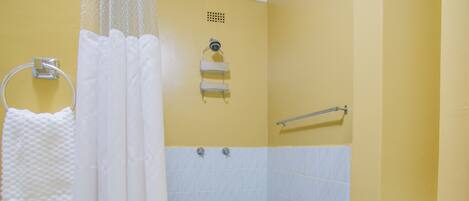 A small, yellow-walled bathroom with a white shower curtain, a towel rack, a handrail, and a tiled floor.