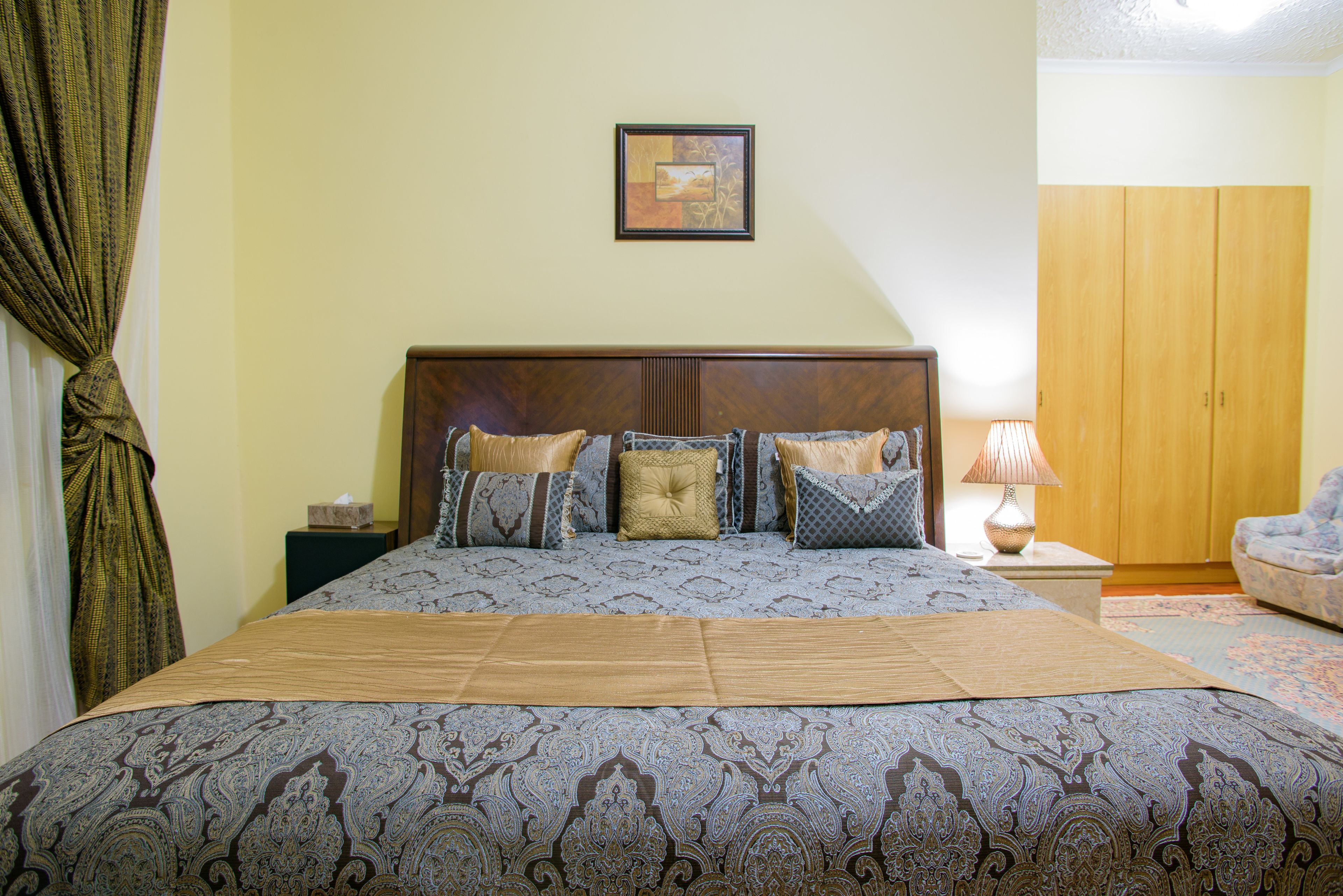 Een net opgemaakt bed met een bedsprei en kussens, een nachtkastje met een lamp, en een ingelijst beeld aan de muur.