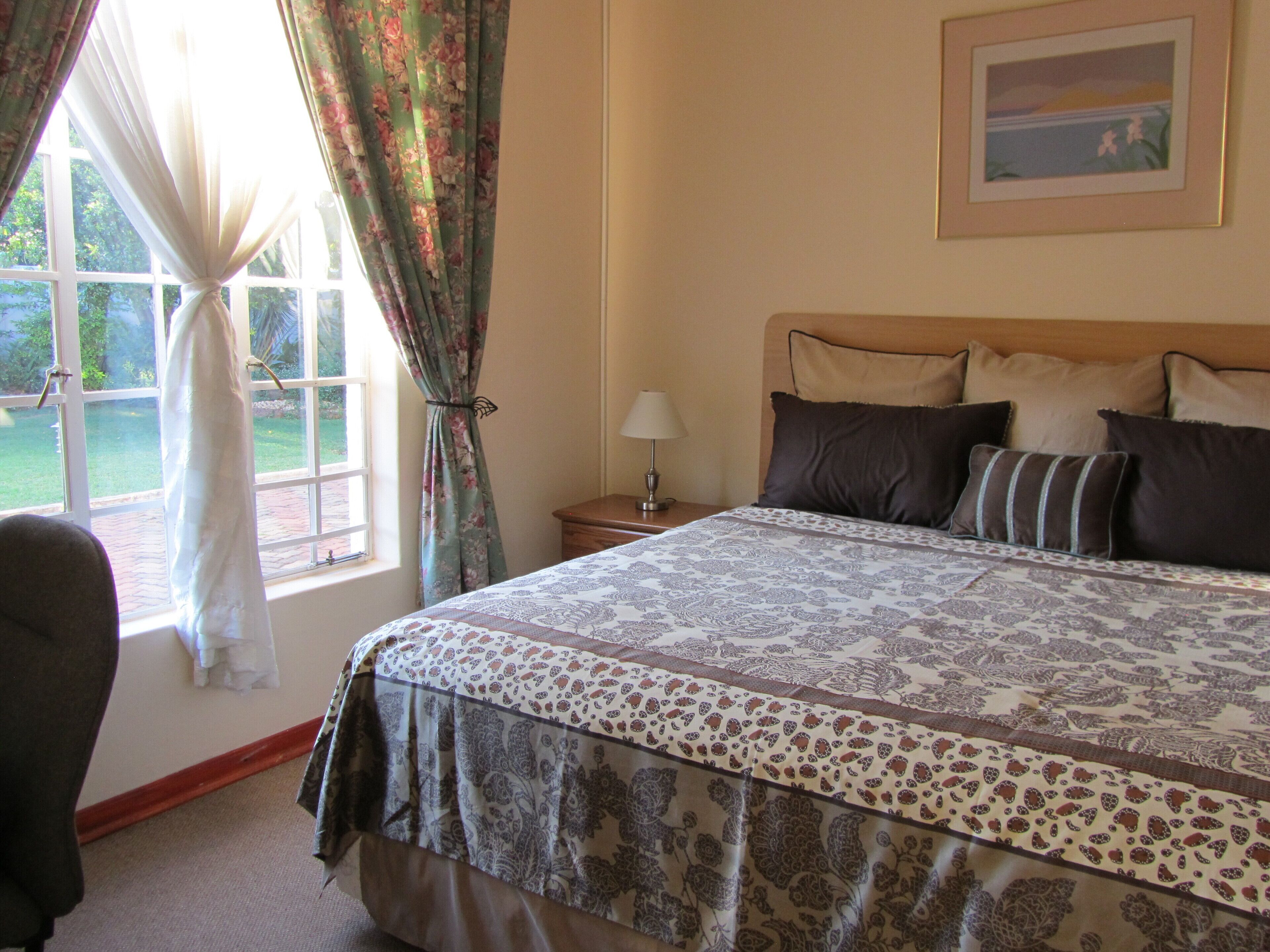A bedroom with a large bed, a nightstand with a lamp, a chair, a window with curtains, and a framed picture on the wall.