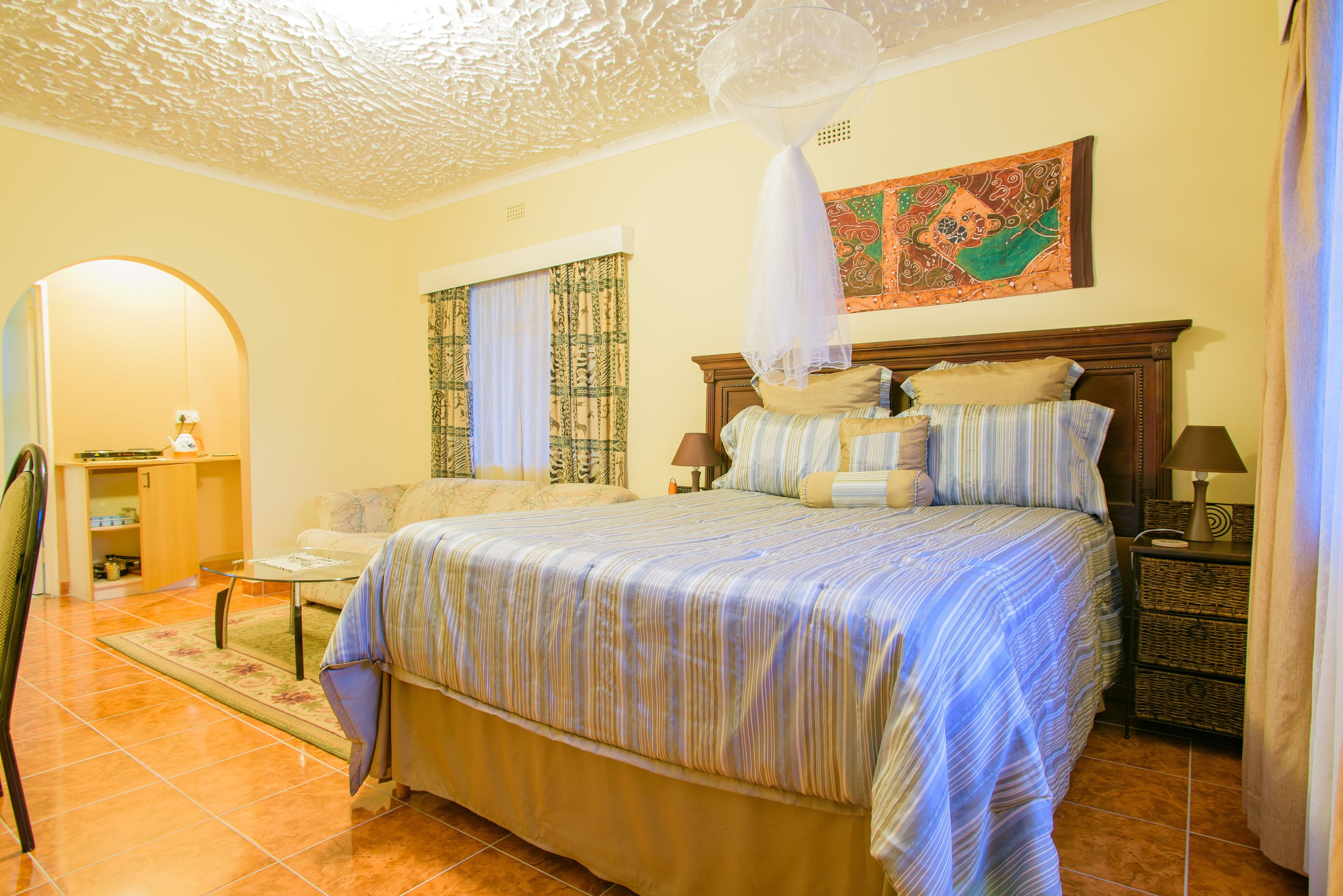 A bedroom with a large bed, a nightstand, a lamp, a chair, a small table, and a decorative ceiling.