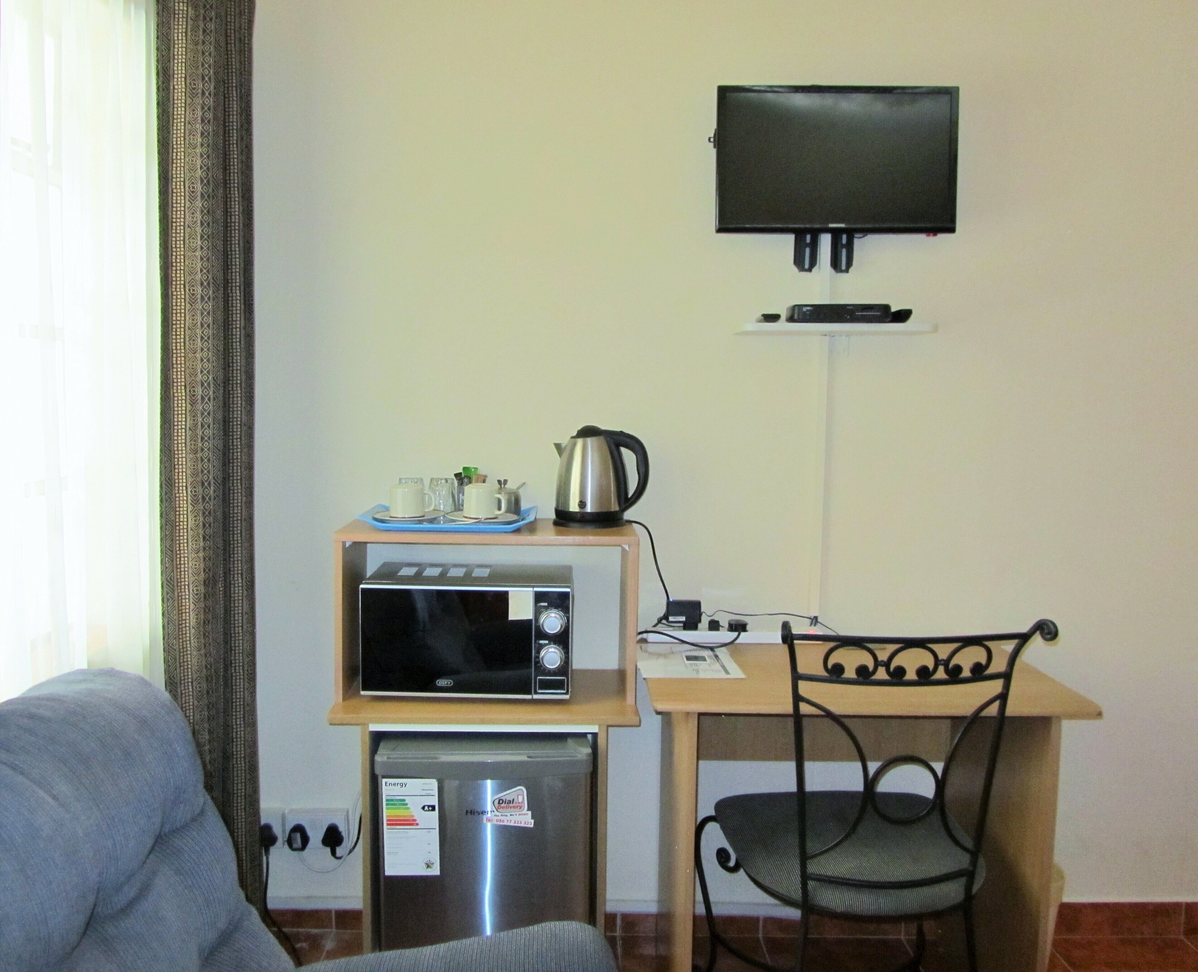 Una kitchenette compacta con microondas, refrigerador y una pequeña mesa con tetera y tazas. Un televisor está montado en la pared sobre la mesa.