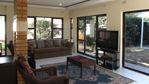 A living room with a sofa, a television, a coffee table, and a chair. There is a large window and a door leading to an outdoor area.