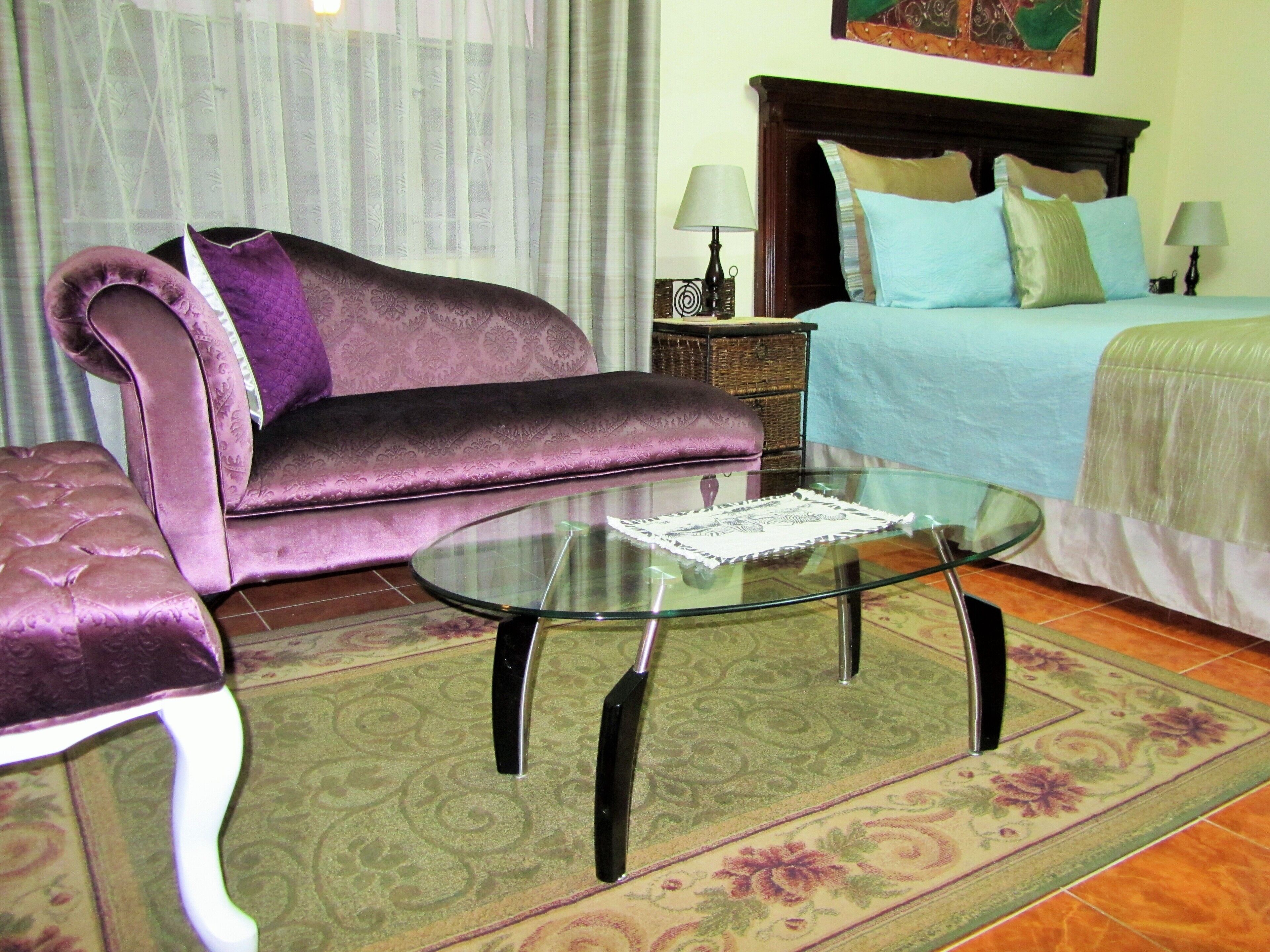 A room with a purple sofa, a glass coffee table, a bed with a blue and green bedspread, and a patterned rug.