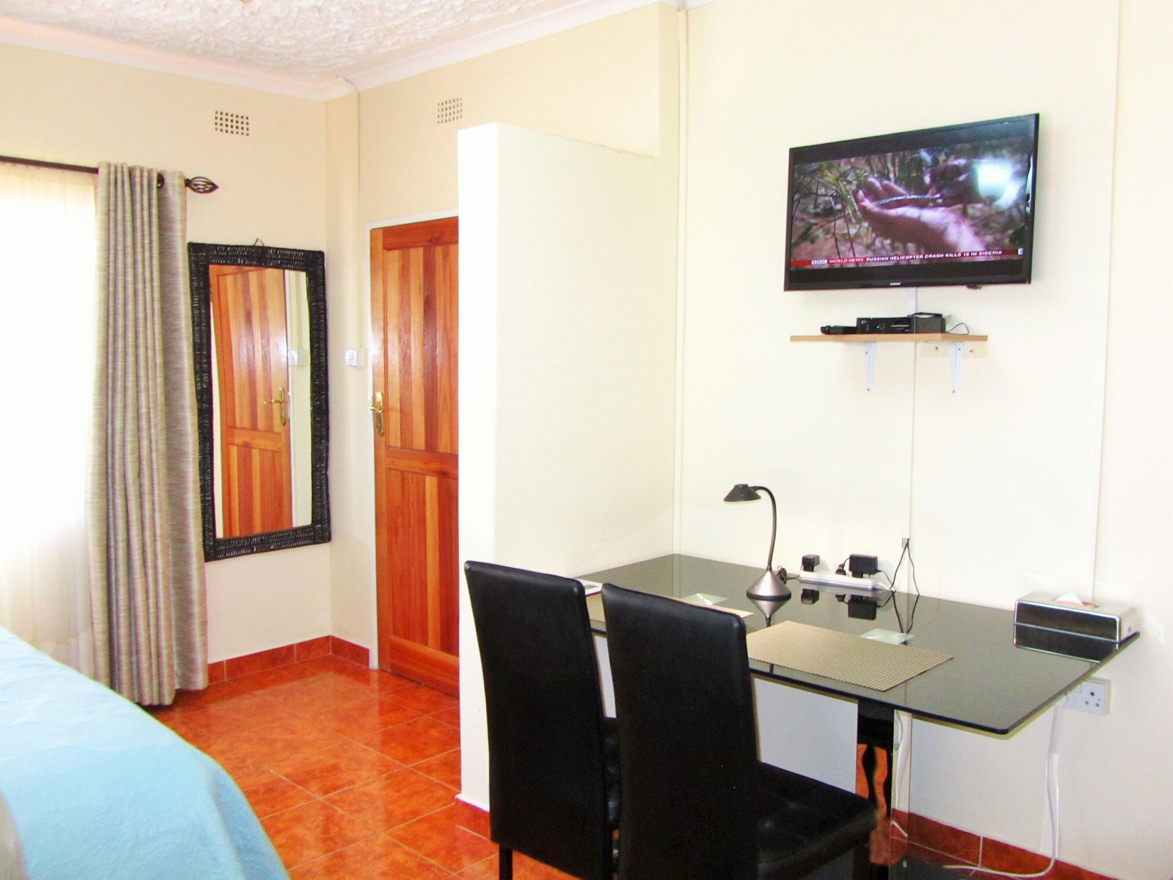 A hotel room with a bed, a desk with two chairs, a television mounted on the wall, and a door.