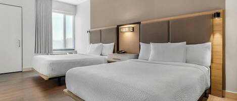 A hotel room with two beds, a nightstand, a bedside table, and a window with blinds.