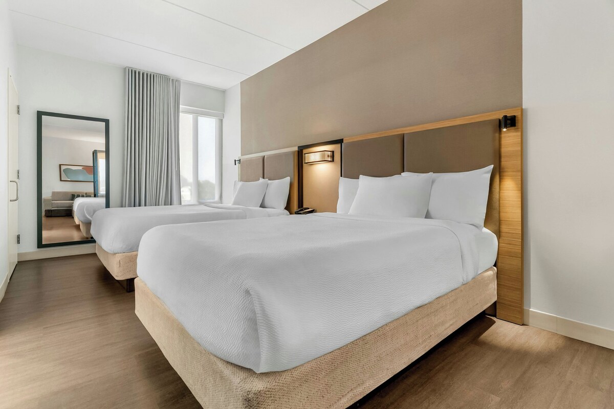 Een hotelkamer met twee bedden, een groot raam met gordijnen, een spiegel en een wandlamp.