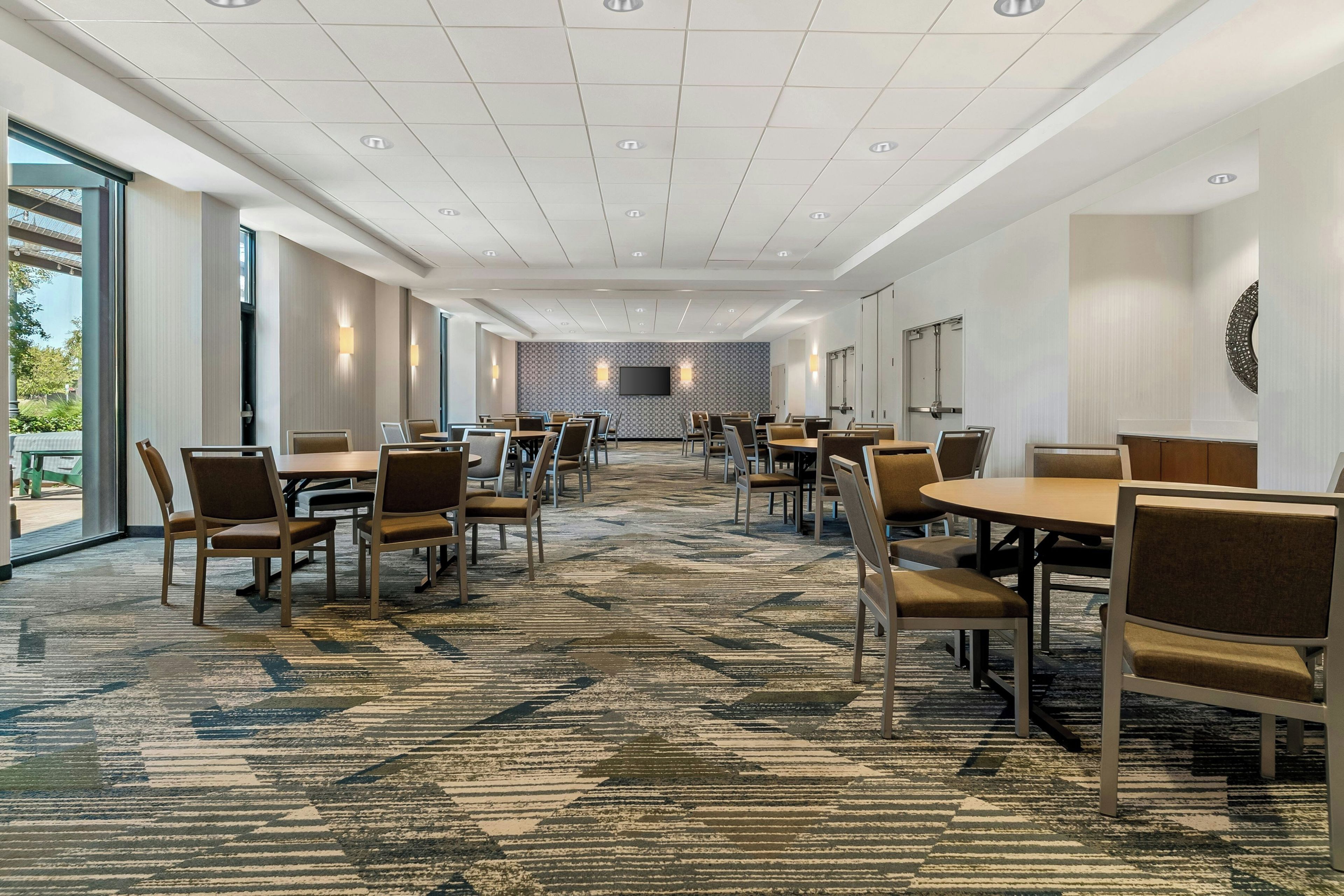A hotel conference room with multiple tables and chairs, a patterned carpet, and a large flat-screen TV on the wall.