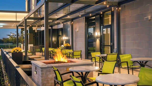 An outdoor patio area with round tables and chairs, a fire feature, and a view of the building's interior. - SpringHill Suites by Marriott Gainesville Haymarket (Gainesville)