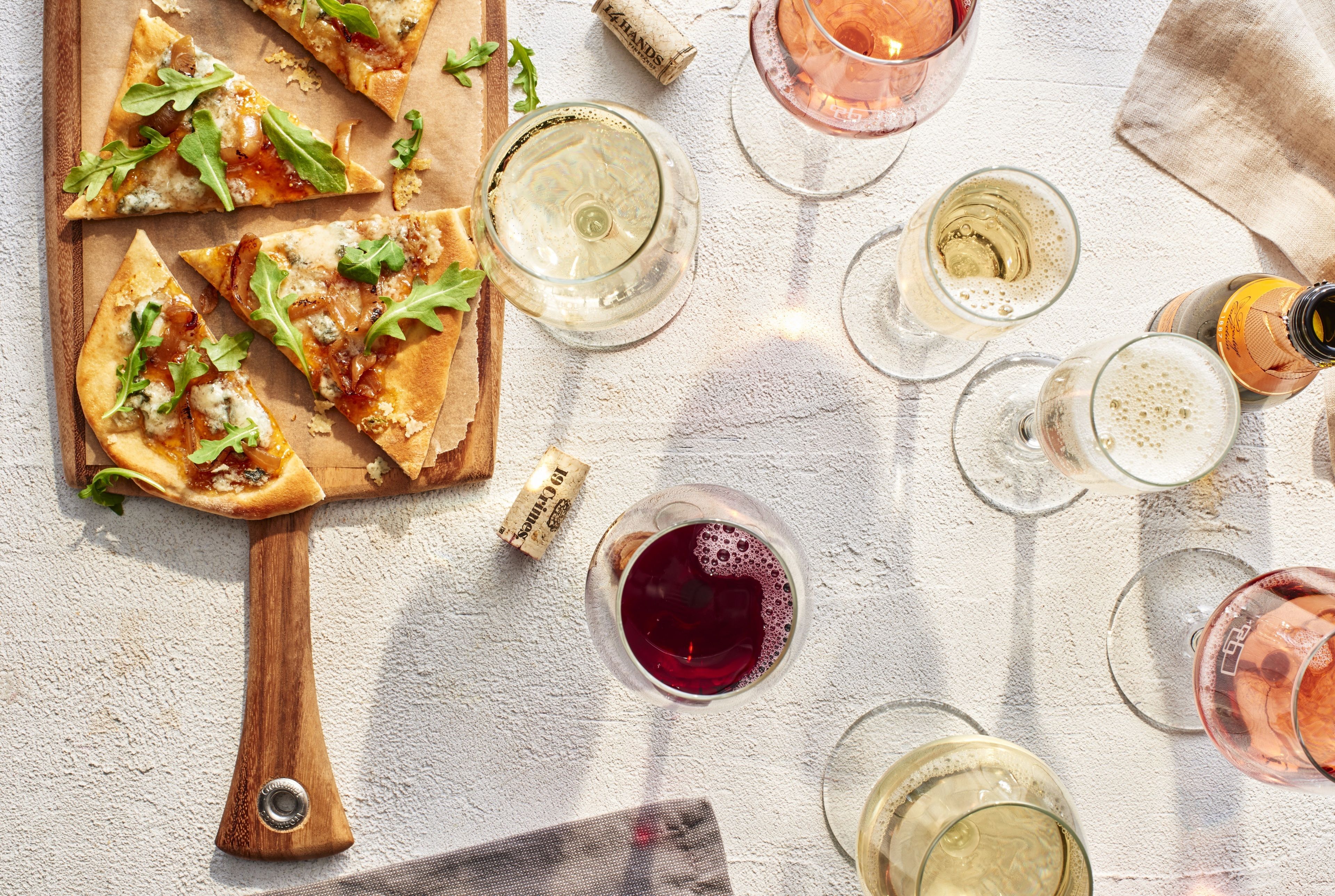 Une planche à pizza en bois, avec des parts de pizza, des verres à vin et un tire-bouchon.