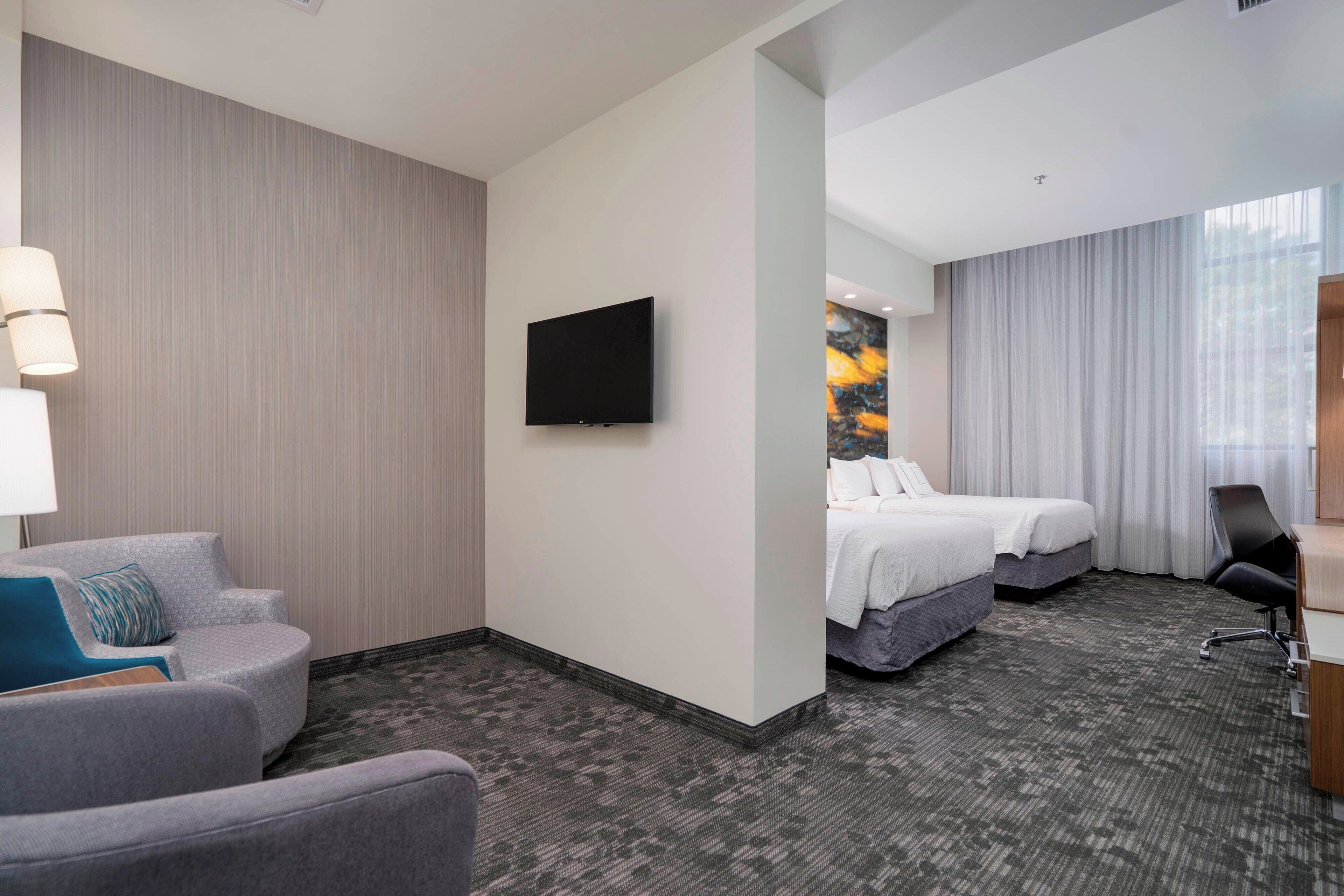 Ein Hotelzimmer mit Flachbildfernseher, zwei Betten, einem grauen Sofa, einem Schreibtisch mit Stuhl und einem großen Fenster mit Vorhängen.