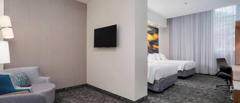 Ein Hotelzimmer mit Flachbildfernseher, zwei Betten, einem grauen Sofa, einem Schreibtisch mit Stuhl und einem großen Fenster mit Vorhängen.