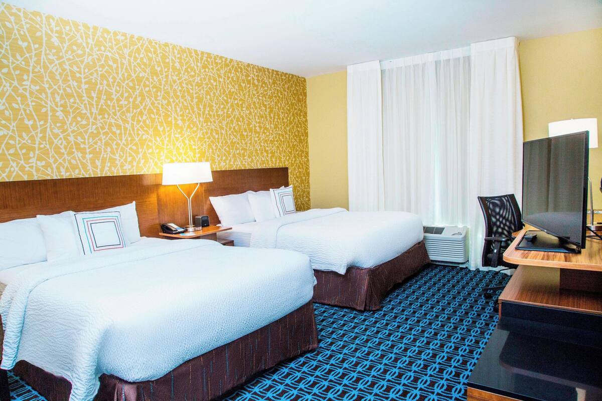 Habitación de hotel con dos camas, un escritorio con televisor y alfombra estampada.