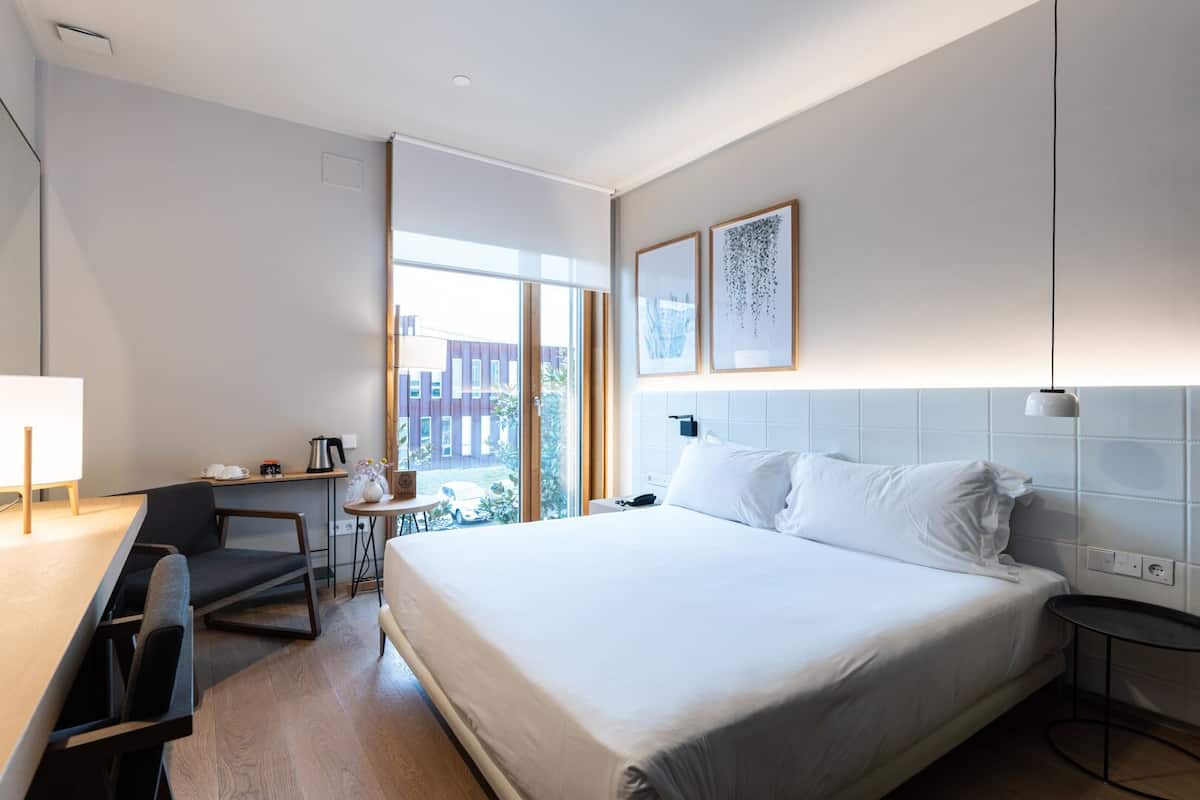 A modern hotel room with a large bed, a desk, and a chair. There is a window with a view of the outside.