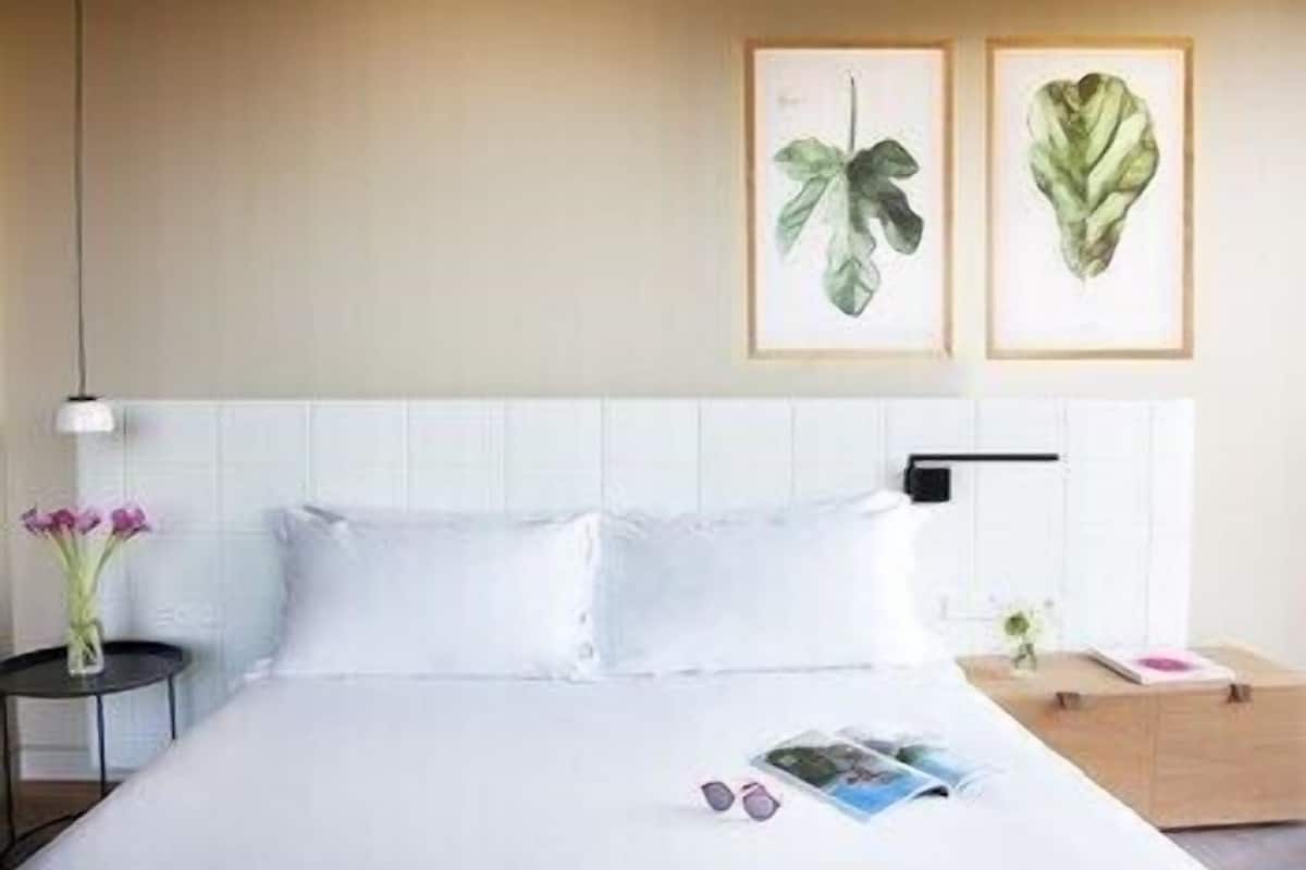 A hotel room with a bed, a bedside table, a nightstand, a lamp, and two framed botanical prints on the wall.