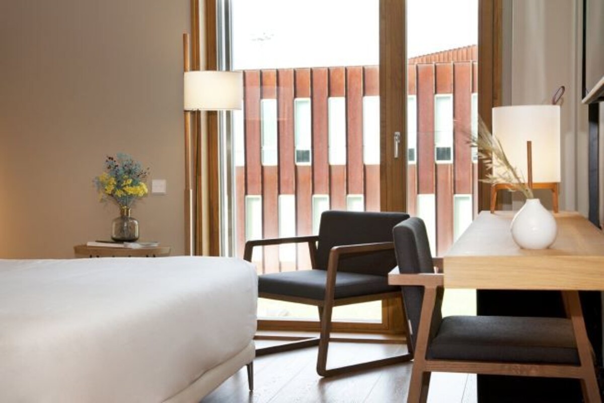 A hotel room with a bed, a desk with a lamp, a chair, and a view of a building through a window.