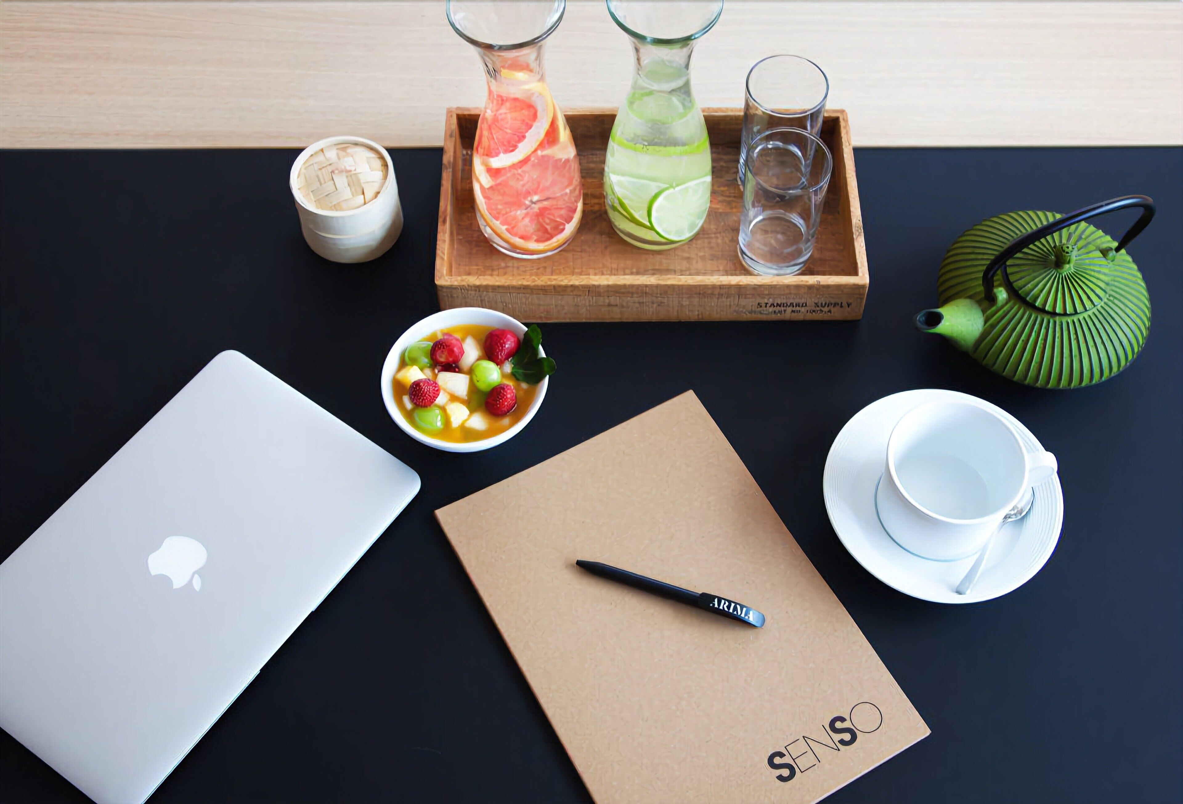 Un espace de travail comprenant un ordinateur portable, un carnet, une théière, une tasse et deux bouteilles en verre contenant des tranches de fruits.