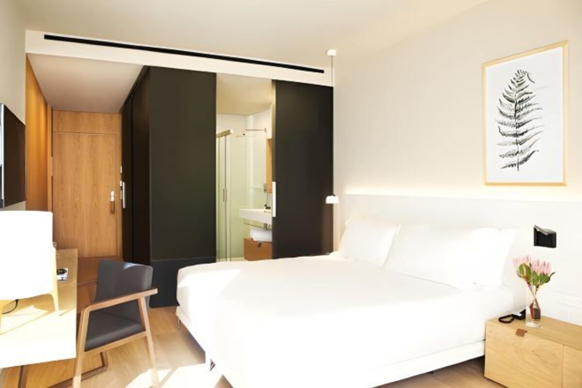 A hotel room with a bed, a desk, a chair, a nightstand, and a framed artwork on the wall.