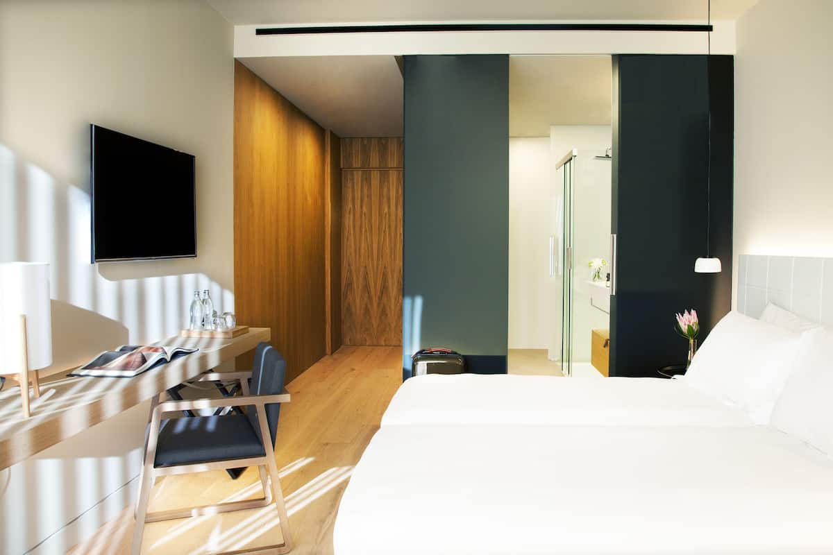 A modern hotel room with a bed, a desk with a chair, a TV mounted on the wall, and a shower area.