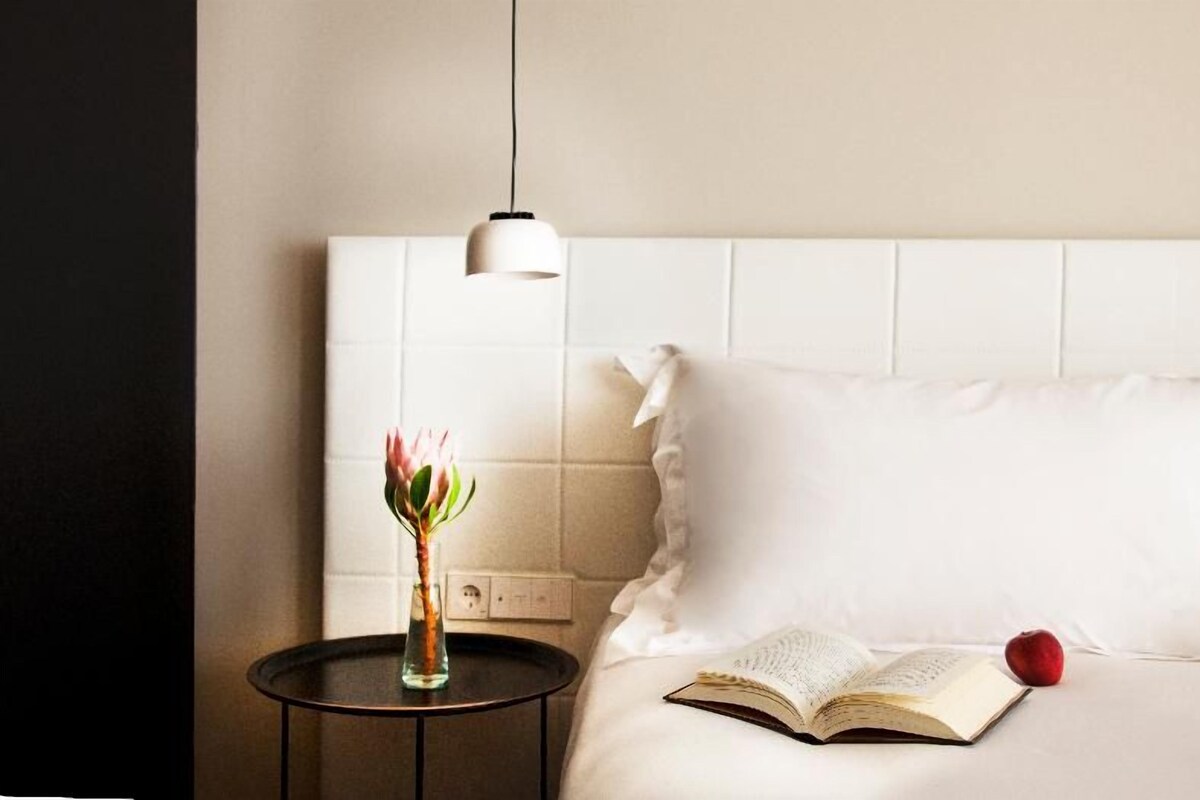 Una cama bien hecha con un libro y una manzana roja a su lado, un florero con una flor en una mesita de noche, y una lámpara colgante.