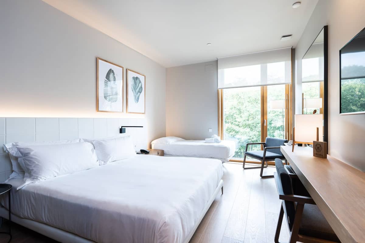 A modern hotel room with two beds, a desk, and large windows offering a view of greenery.
