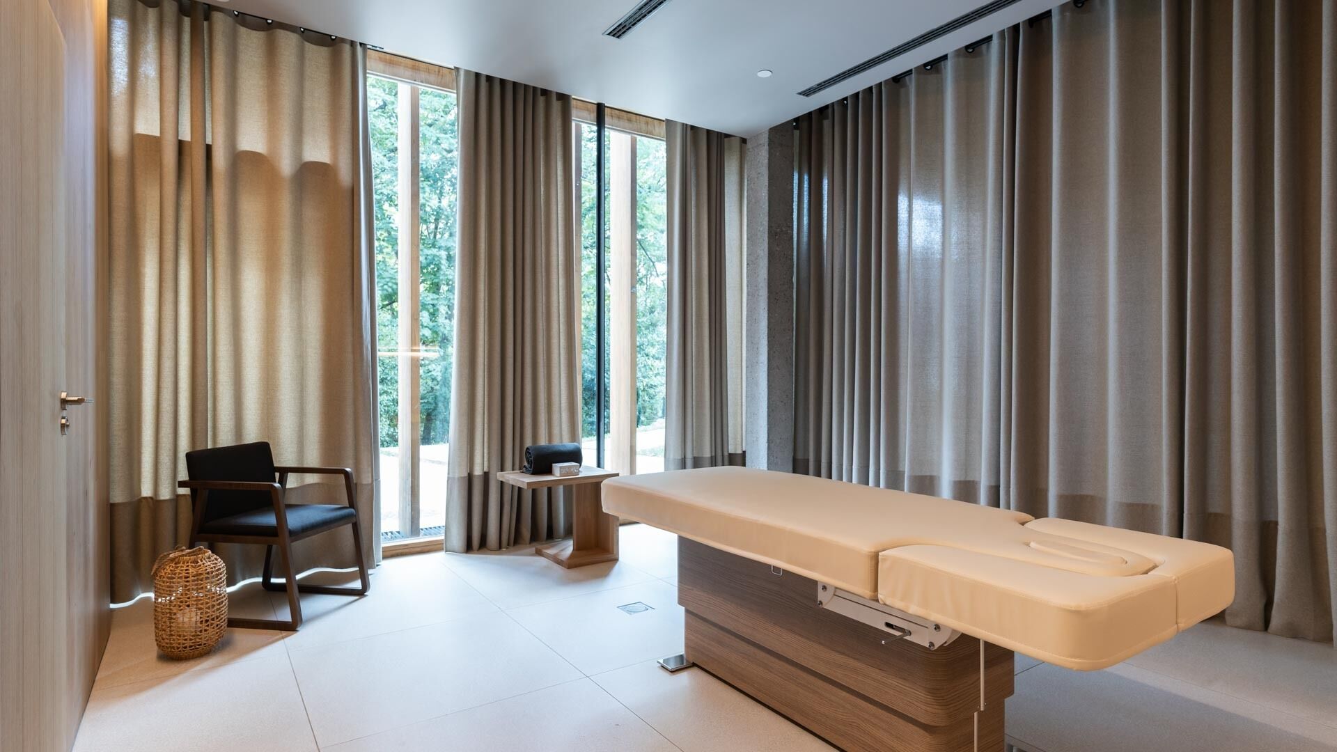 Une pièce moderne équipée d’une table de massage, d’une chaise, d’une petite table et d’une fenêtre avec des rideaux.