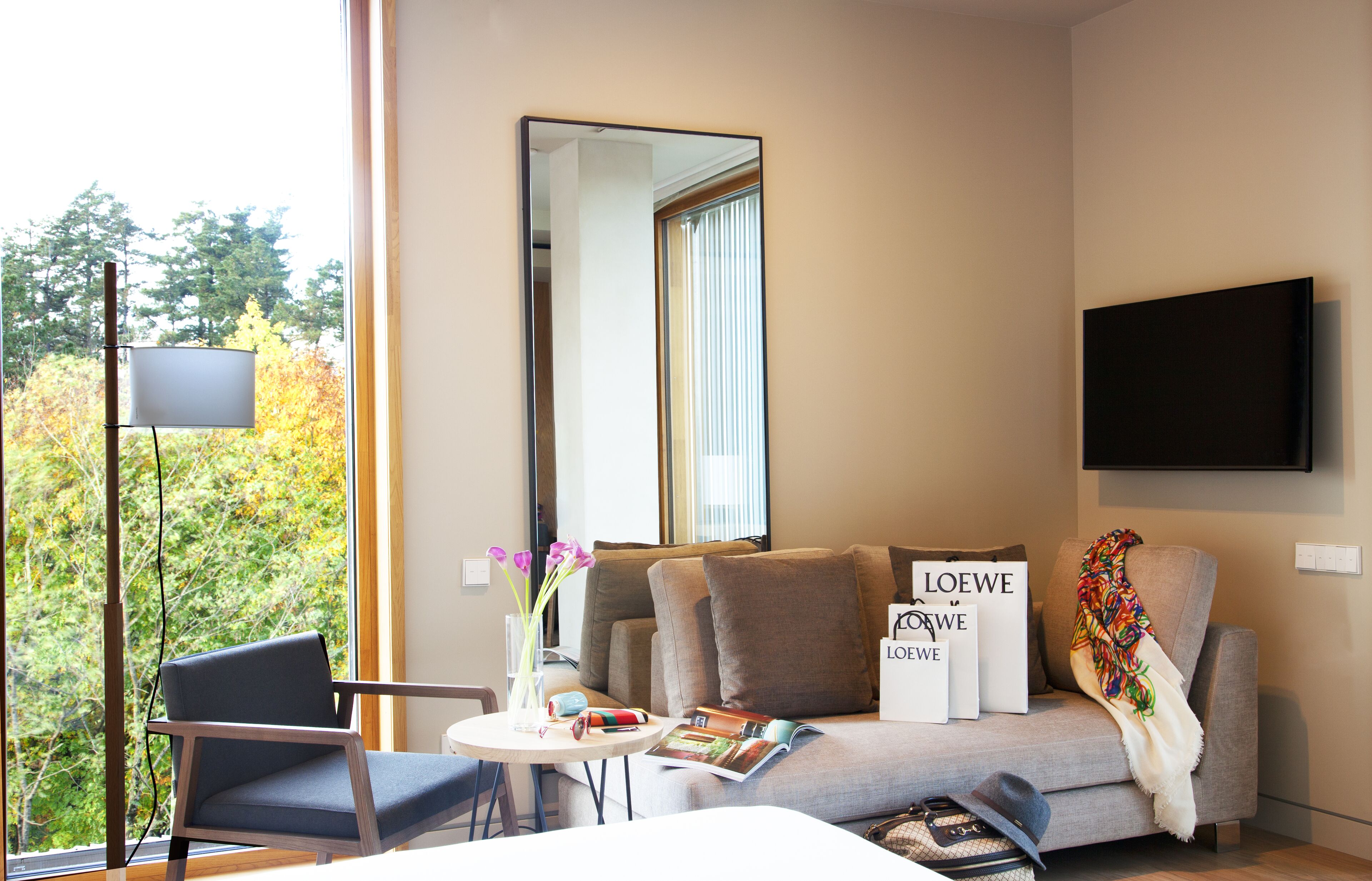 Una habitación de hotel moderna con sofá, una mesita con un jarrón de flores, una silla, un televisor de pantalla plana y un ventanal con vistas a árboles.
