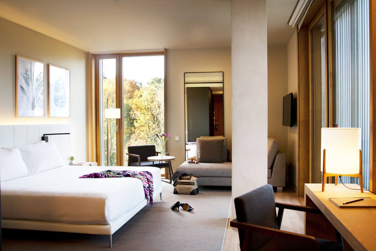 A hotel room with a bed, a desk, a chair, a TV, and a window with a view of trees.