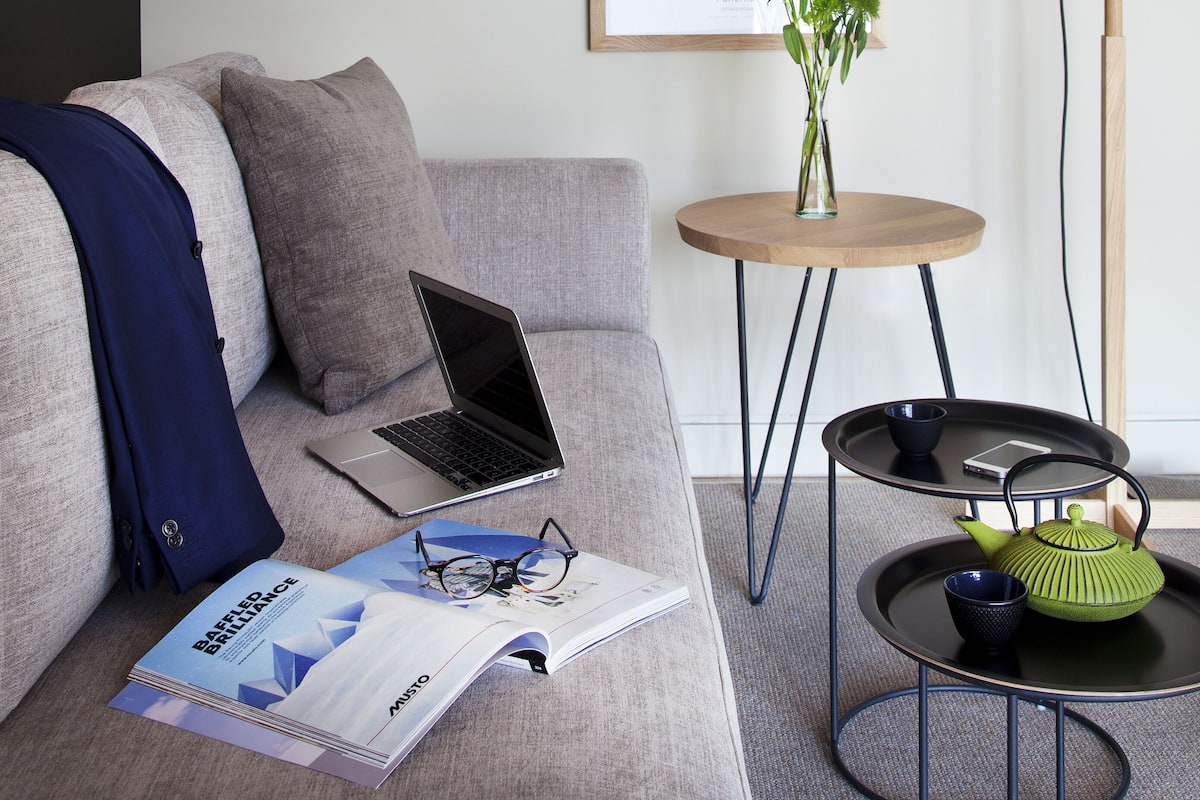 A modern living room with a grey sofa, a laptop, and a magazine. There are framed artworks on the wall, a small round table with a vase of flowers, and a floor lamp.