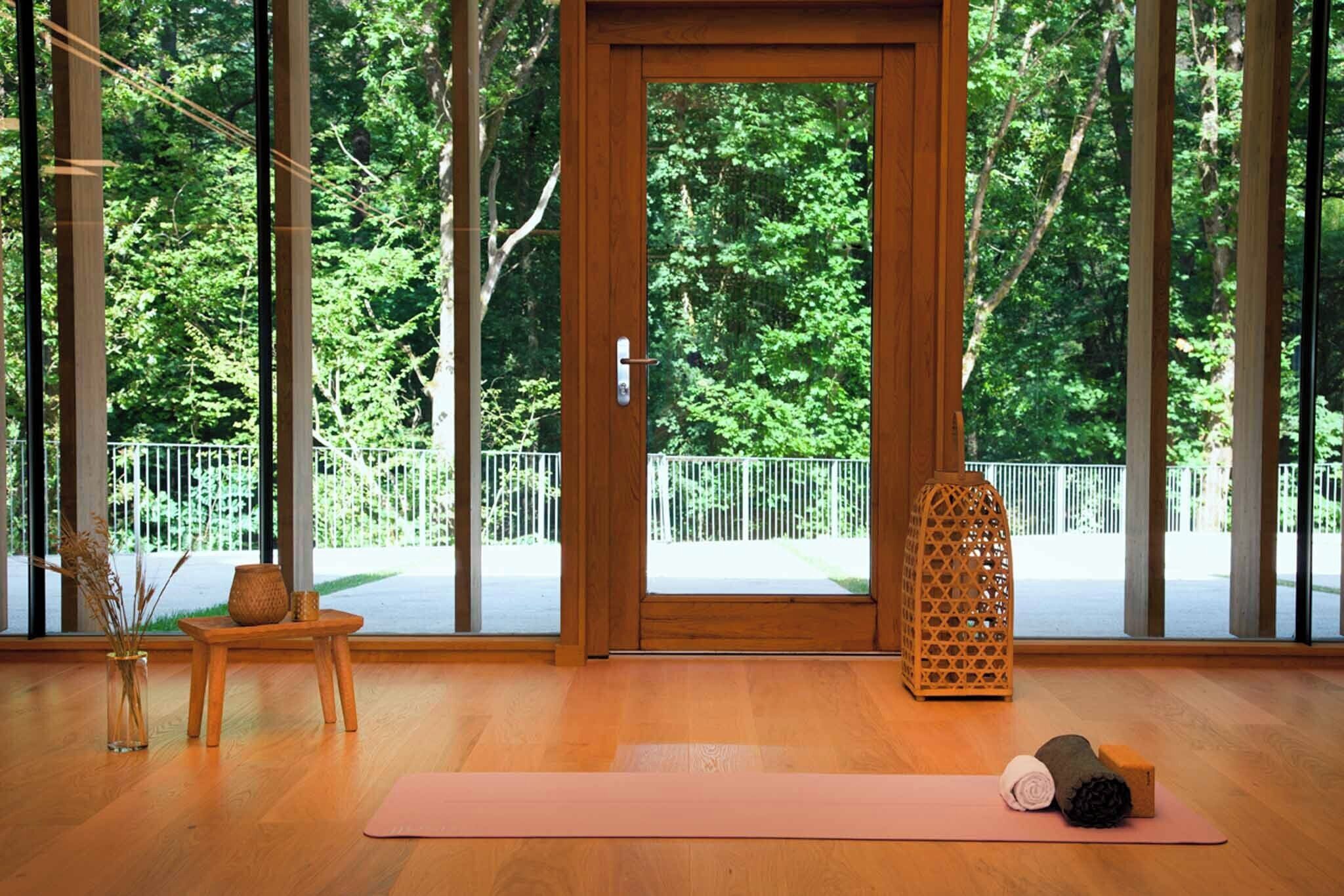 A room with a wooden floor, a yoga mat, a small wooden table with a vase and candle, and a large window with a view of trees and a fence.
