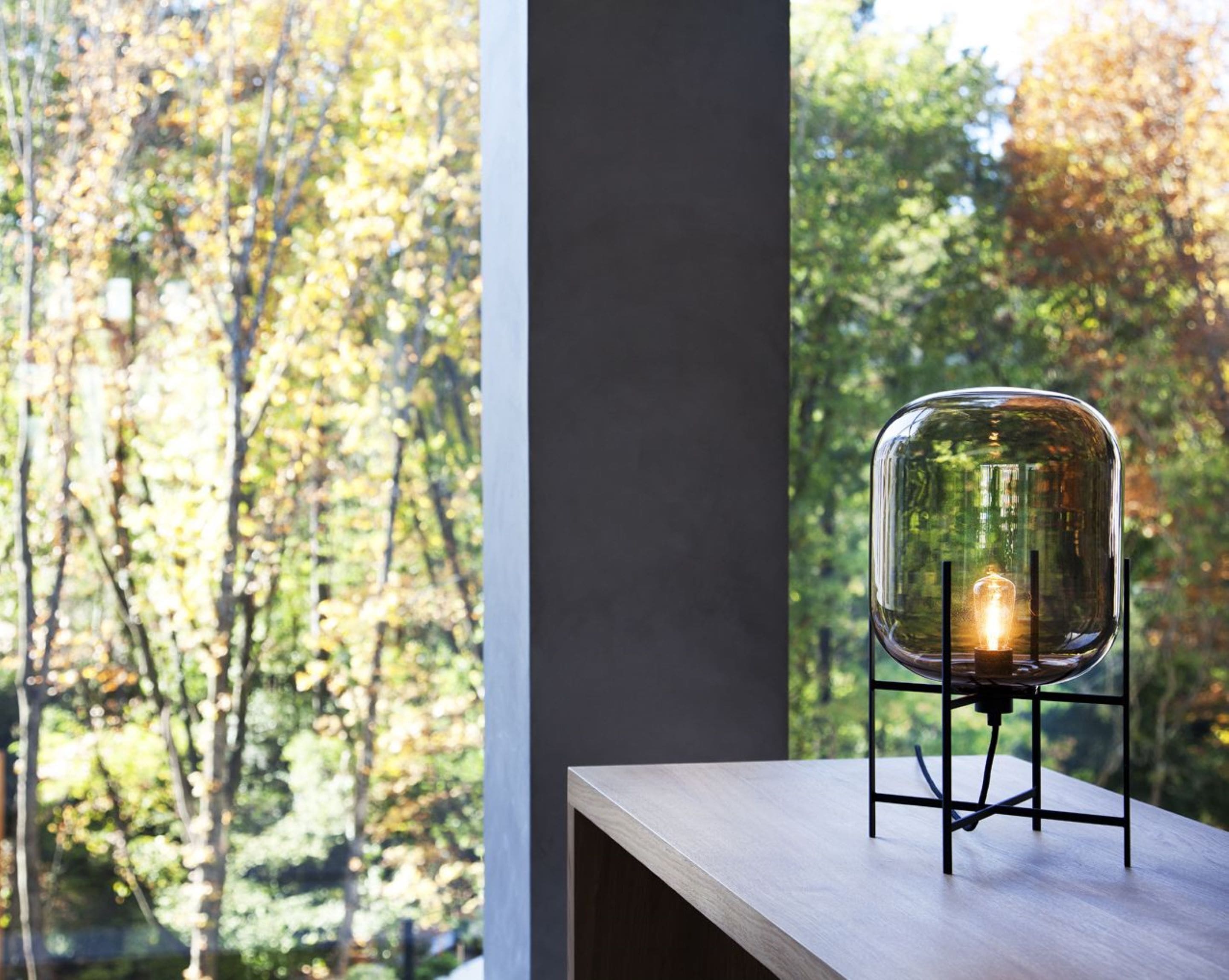 A modern chair with a glass enclosure and a light bulb inside, placed on a wooden table by a window with a view of trees.